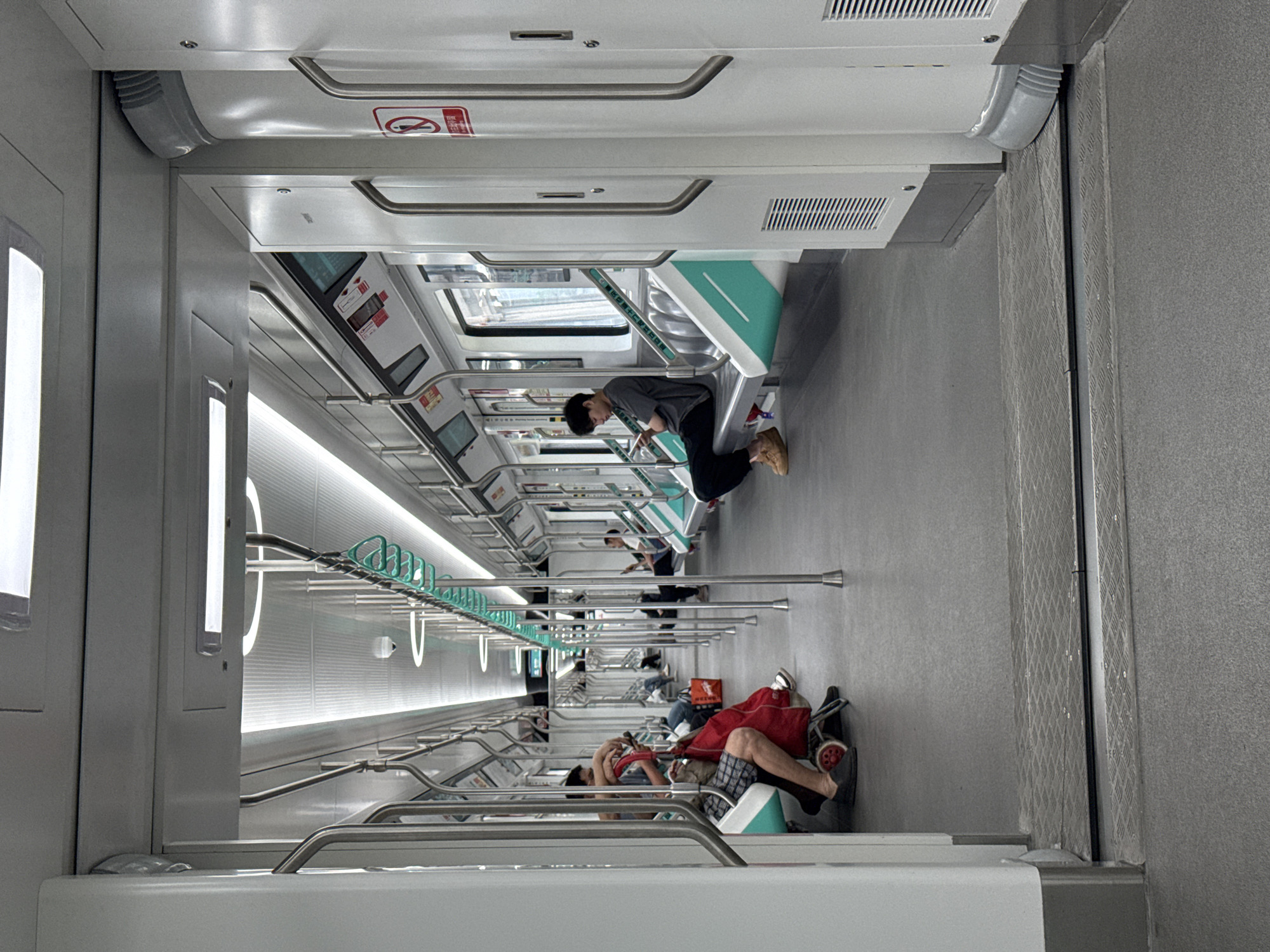 The image, initially rotated 90 degrees clockwise, depicts the brightly lit interior of a modern subway or train car, likely in Tiaodeng, China, as indicated by the context. The train car features a clean aesthetic dominated by white and light grey surfaces, accented with a distinct mint green on the seating and some fixtures.
The scene shows several passengers engaged in travel. In the mid-ground, a young person wearing a grey top and dark pants is seated, looking downwards, possibly at a phone or book. Further into the background, other passengers are visible, some seated and some standing, though their details are less clear. In the foreground, partially obscured, a person is seated with their legs extended, appearing to rest or sleep, with a red bag or trolley next to them. The overall atmosphere appears calm and unhurried.
The interior is well-appointed with numerous stainless steel handrails running along the ceiling and vertically between seating areas, along with mint green hanging straps for standing passengers. Long, linear LED light strips illuminate the ceiling and upper walls, contributing to the bright environment. The floor is light grey, with a textured, possibly non-slip, surface, and a section near a door features a diamond-plate metallic pattern. Overhead, white panels integrate air vents.
A notable detail is a red sign with white Chinese characters visible above a seat, which reads "禁止倚靠车门" (Jìnzhǐ yǐkào chēmén), meaning "No leaning on the car door." This text confirms the location within a Chinese-speaking region and reinforces its identity as a public transport vehicle. The car design, including the specific color scheme and modern fixtures, is characteristic of contemporary subway systems.