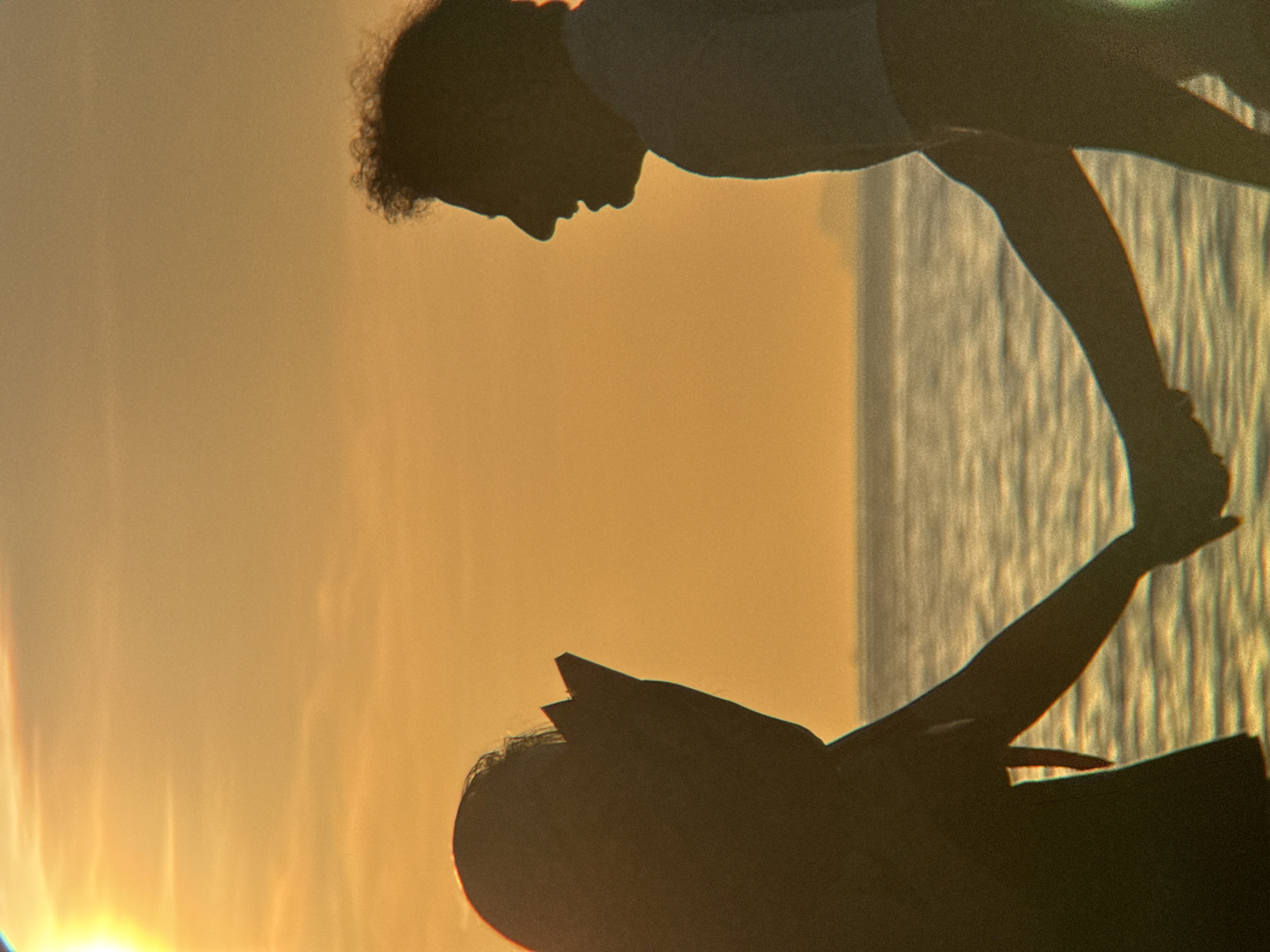 A silhouette of a couple holding hands stands on a beach in Nha Trang, Vietnam, during sunset. The sky is a warm orange, with the sun positioned in the upper left corner. The sea stretches out behind them, with gentle waves reflecting the sunset light. The couple is facing each other, with the woman on the left and the man on the right. The woman appears to be wearing a crown or headpiece. Their bodies are mostly obscured by shadow, but their connected hands are clearly visible, suggesting a moment of affection. The overall scene evokes a romantic and peaceful atmosphere.