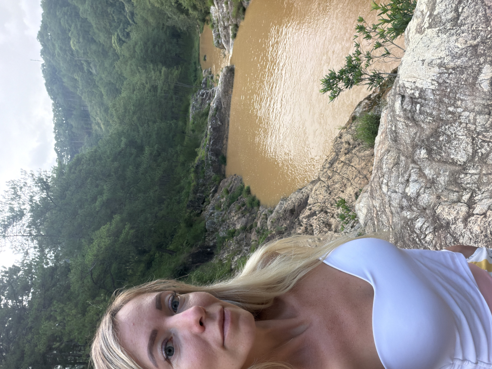 A blonde woman with blue eyes, wearing a white strapless top and patterned shorts, stands on rocks overlooking a muddy brown river. She is facing the camera, with the river and a densely forested hillside in the background. The location is identified as Păng Tiêng 1, Vietnam.