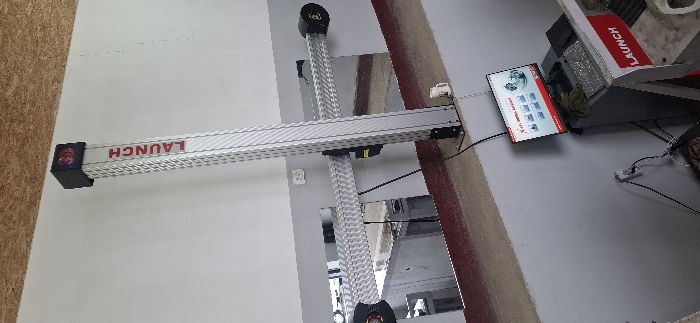 An automotive wheel alignment system by Launch is positioned within a garage. The system features a horizontal bar with "LAUNCH" printed on it, supported by two vertical poles. A computer console with a display screen is located to the right of the equipment. The console's display presents various vehicle-related data and graphics. A power outlet is visible on the wall.