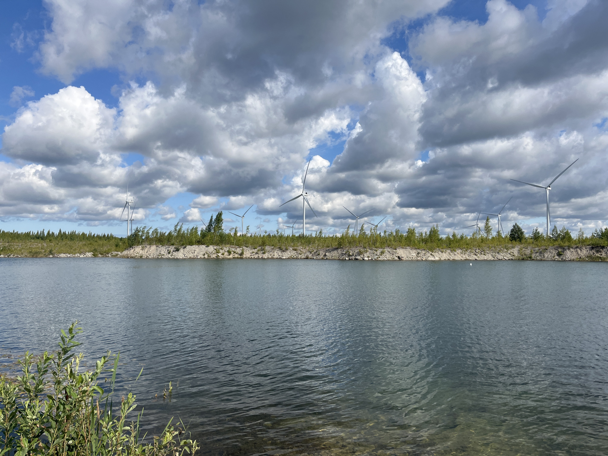 In Ojamaa, Estonia, a body of water with visible rocks on the bottom is in the foreground. The water's surface is rippled, reflecting the sky. Behind the water, a strip of land is covered with green trees and some lighter colored rocky areas. In the background, several white wind turbines stand against a sky filled with a mixture of white and gray clouds. The scene is captured under daylight.