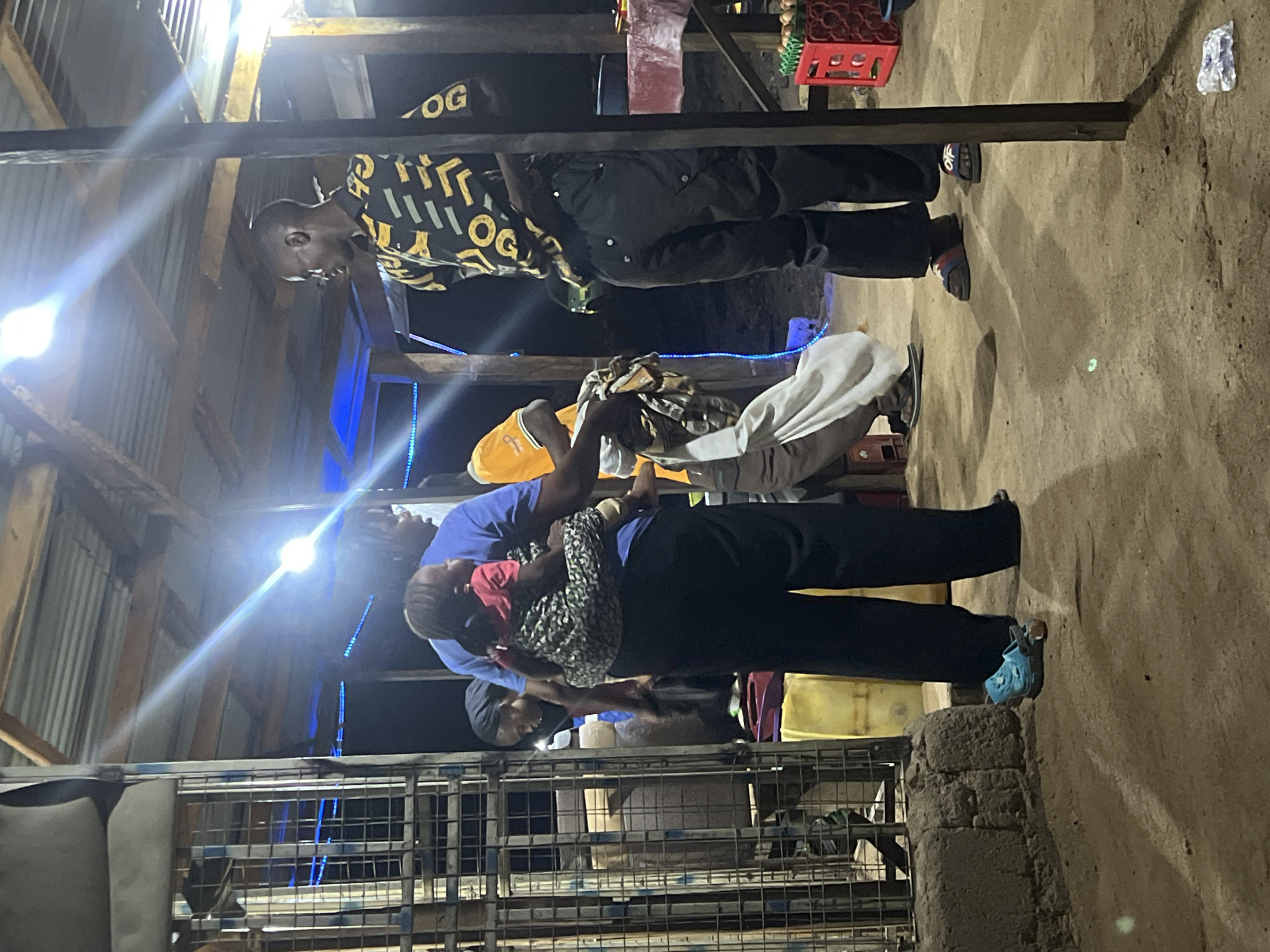 The image appears to be taken indoors in Jalingo, Nigeria. Several people are present, including a person holding a small child. Another person is standing next to them. One person is wearing a yellow and black patterned shirt. The setting includes corrugated metal walls and support beams. Light sources are visible in the scene. The floor is a light-colored earth.