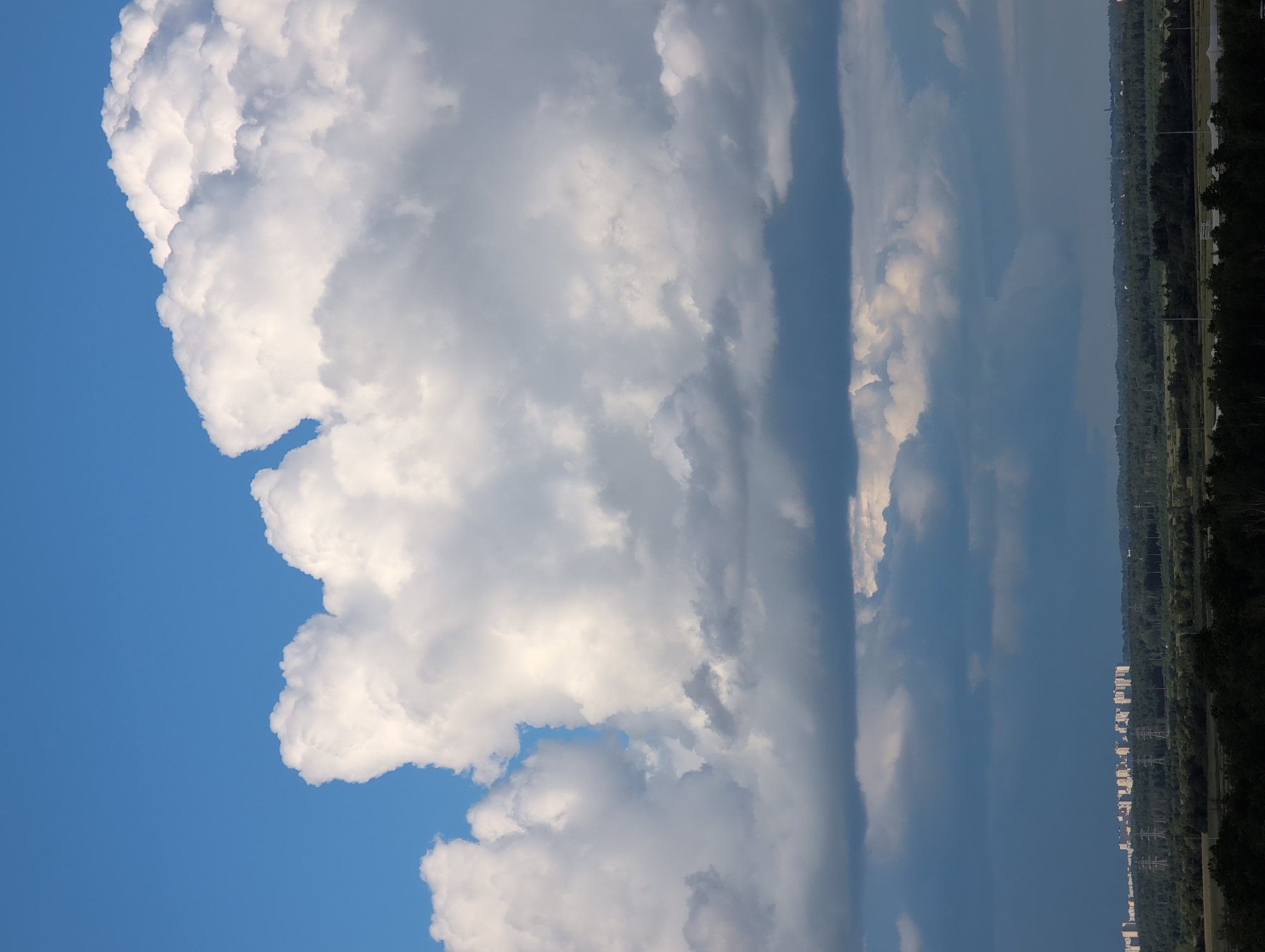 This is a vertical, eye-level, medium shot of a vast sky with dramatic clouds and a distant cityscape. The dominant subject is the sky, filled with a large, fluffy white cumulus cloud formation that takes up the left side of the frame. Its textured surface and voluminous shape suggest a bright, perhaps slightly humid day. To the right of the cloud, the sky transitions to a lighter blue, with streaks of darker, more diffused clouds.
In the far distance, along the right edge of the frame, a strip of land is visible. This appears to be a suburban or semi-urban area with buildings and lush green trees. The buildings are predominantly white and arranged in a somewhat linear fashion, suggesting a planned community or development. There are also visible power lines and streetlights, indicating infrastructure. The overall impression of the cityscape is one of order and a connection to nature with the abundance of greenery.
The image appears to have been taken during the daytime, likely late afternoon or early evening, given the quality of the light. The bright white of the clouds and the blue of the sky suggest good weather, possibly with some cloud cover contributing to the dramatic sky. The location is identified as Saint Petersburg, Russia. No people or specific objects are clearly discernible, focusing the attention on the natural elements of the sky and the distant man-made landscape. There is no visible text in the image.
