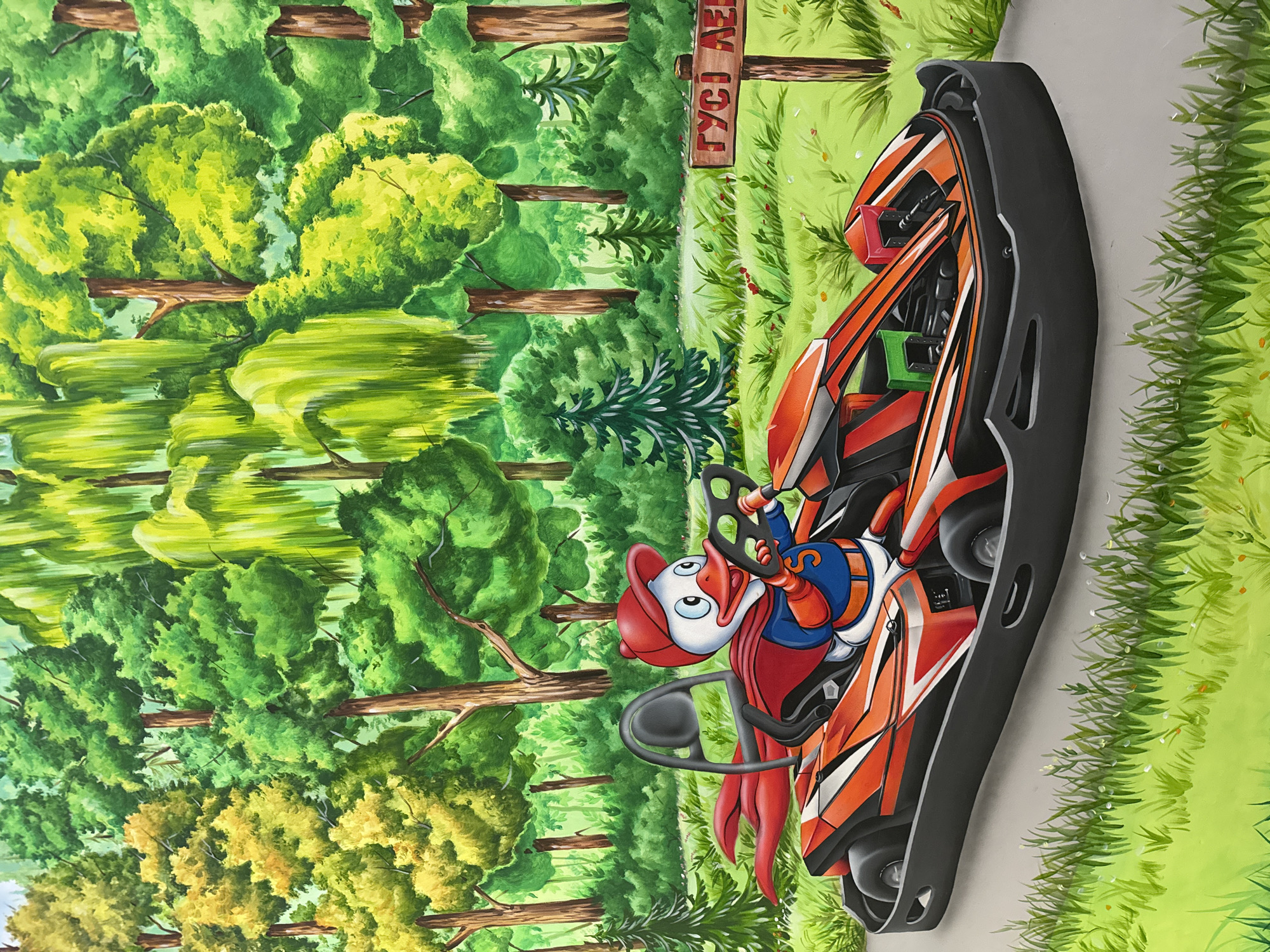 This is a brightly colored, stylized illustration of a cartoon duck character driving a go-kart through a forest. The duck is wearing a red cap, a red cape, and a blue and yellow superhero-like uniform with a red "S" emblem on the chest. The go-kart is orange and black with white accents. The background depicts a vibrant forest with lush green trees of various types, some with yellow foliage, and a dappled sunlight effect. In the foreground, there is a grassy area with small red flowers. To the right of the go-kart, a wooden signpost with Ukrainian text is visible. The scene is playful and whimsical. There is no indication of this scene being located in the city of Orlivshchyna, Ukraine.