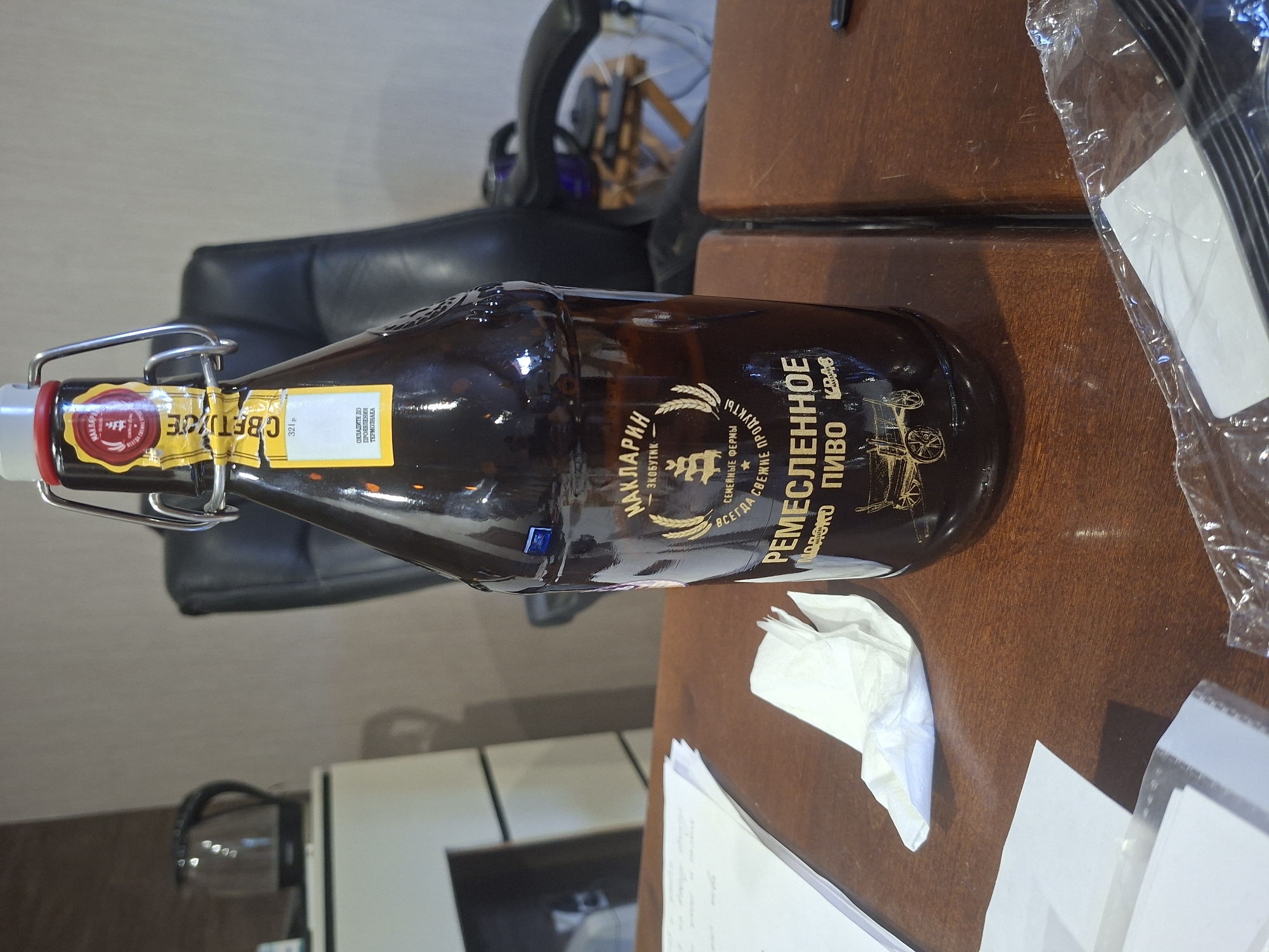 A flip-top glass bottle of dark beer is situated on a wooden table in what appears to be an office or living space. The bottle has a label with Cyrillic text. To the left of the bottle, a stack of white papers and a crumpled white napkin are visible. In the background, a black leather office chair is partially visible. Further back, a portion of a white cabinet and a dark electric kettle are present. The overall environment suggests an indoor setting. The image does not provide any information or context to determine its location within the city of Ivanovo, Russia.