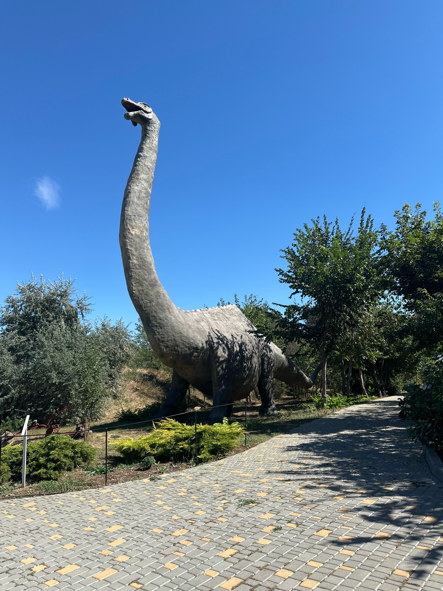 A grey, concrete sculpture of a dinosaur with a long neck and tail stands on a grassy slope under a clear blue sky. The dinosaur is positioned to the left of a paved walkway made of interlocking grey and yellow bricks. Lush green trees and bushes surround the dinosaur and the walkway. The scene appears to be outdoors, possibly in a park or a themed attraction, located in the city of Molodizhne, Ukraine. There are no people visible in the image.