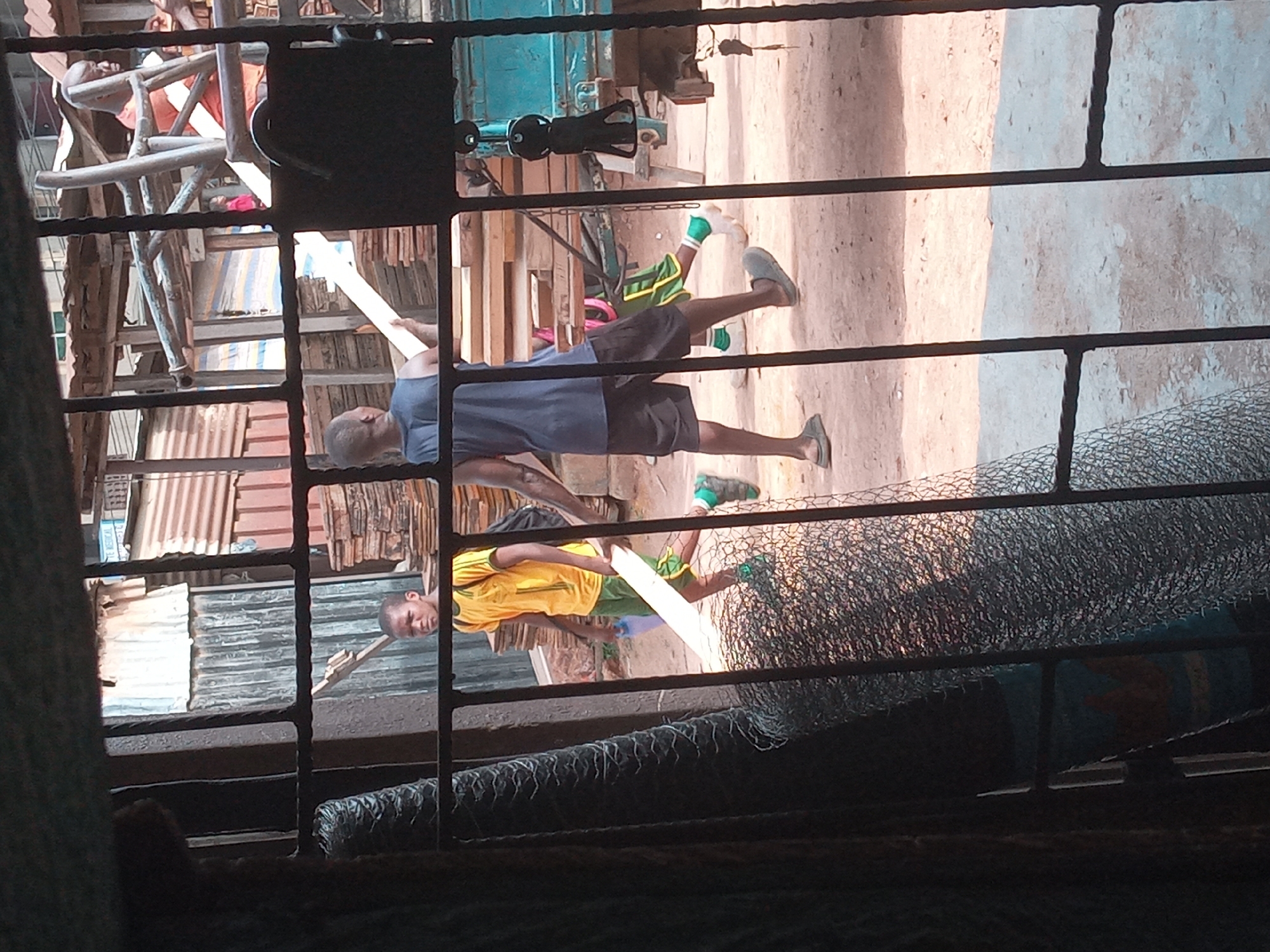 From behind a barred window, the scene is set in Nkpor, Nigeria. Two men are visible outdoors. One man wears a blue shirt and dark shorts, and the other wears a yellow shirt and green shorts. Both men hold long, rectangular, light-colored objects, possibly boards or planks. The environment includes corrugated metal structures and miscellaneous objects in the background, indicating a likely commercial or residential area. Rolls of wire mesh rest behind the bars, further obscuring the view.