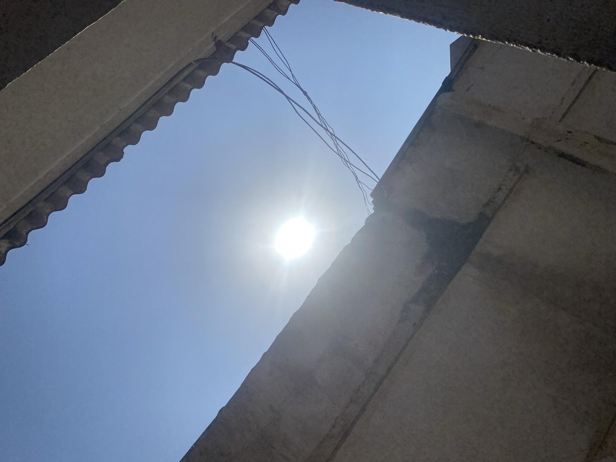 The media file shows a view looking upwards towards a bright, sunlit sky. The sun is prominently featured, appearing as a radiant white orb with visible sun flares. The sky is a clear, vibrant blue.
Framing the sky are elements of what appears to be a partially constructed or unfinished building. On the left, a corrugated surface, possibly roofing material, extends downwards at an angle, with a solid concrete structure above it. On the right, a rough concrete beam or wall occupies the lower right portion of the frame. The concrete surfaces are textured and appear weathered, with some darker patches and lines suggesting age or damage.
Across the sky, several thin black wires are visible, crossing each other in the upper central part of the image. They appear to be suspended from unseen points above.
The strong sunlight suggests it is daytime, likely midday given the intensity and the position of the sun. The clear sky indicates fair weather. The presence of unfinished construction elements and exposed wires might hint at a location in a developing area or an ongoing construction project. The overall impression is one of an outdoor scene, viewed from a confined space within a structure, looking out towards the open sky. There is no visible text in the media file. The scene evokes a sense of bright daylight and the raw, exposed nature of a construction site.
