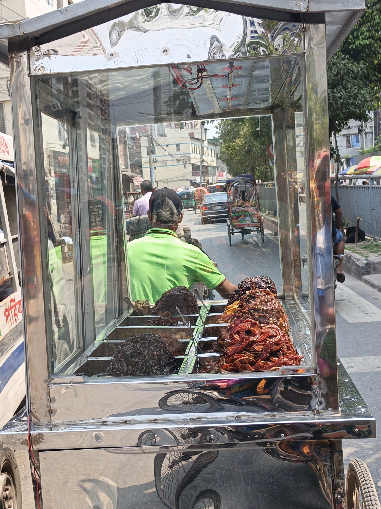 A street food vendor is operating a metal cart on a busy street in Dhaka, Bangladesh. The vendor, wearing a green shirt, is positioned behind the cart which contains a variety of prepared foods, some dark and some red in color. The cart is enclosed in glass on three sides and reflects the surrounding buildings and traffic. In the background, there are several vehicles, including cars and rickshaws, moving along the street, and other pedestrians are visible.