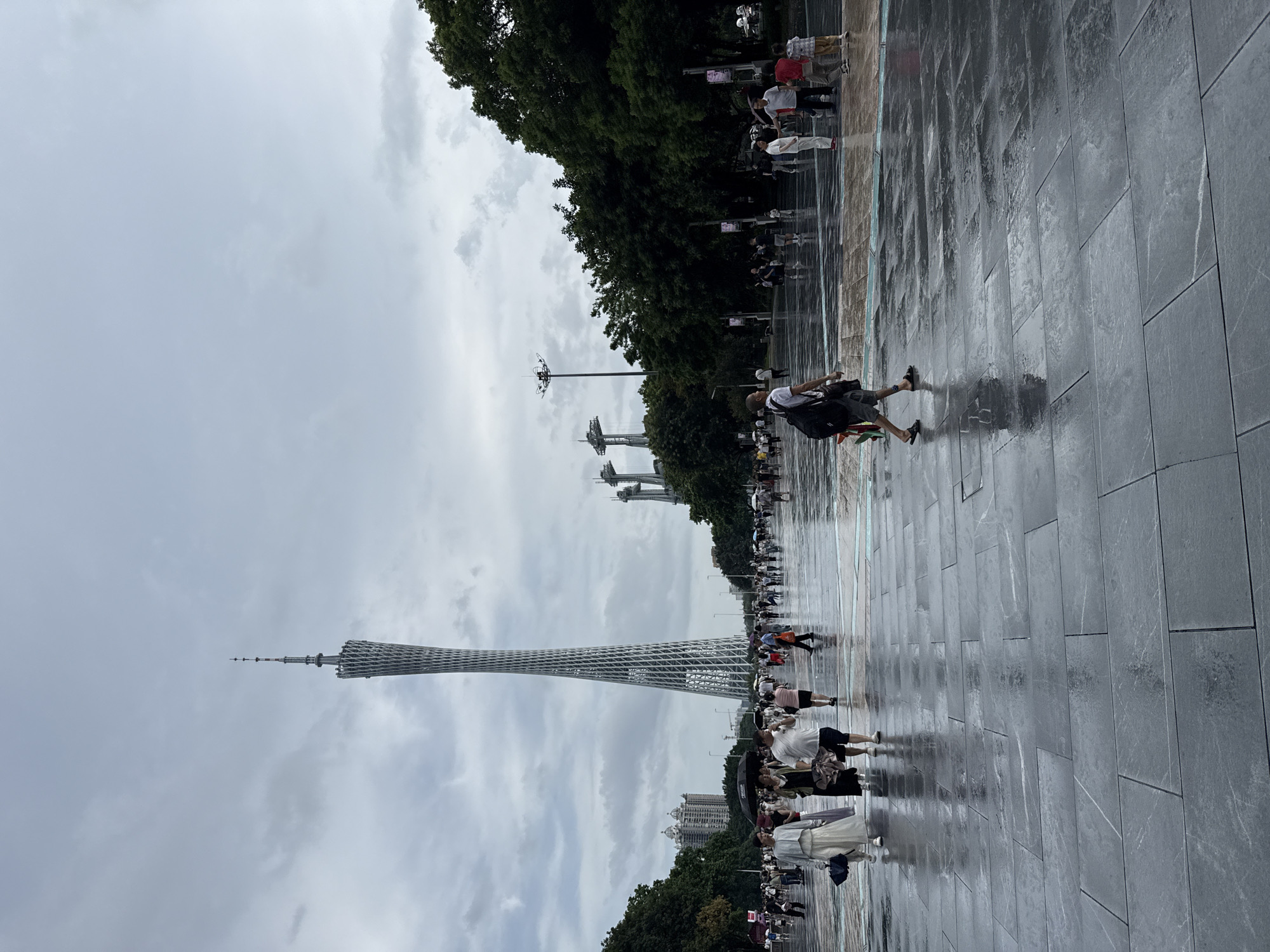 The image shows a wide, wet, paved plaza with several people walking. In the background, a tall, lattice-like tower dominates the skyline. The sky is overcast with grey clouds. To the right of the tower, trees line the edge of the plaza. In the foreground, people are walking in various directions. One man in the foreground is walking away from the camera with a backpack. The ground is wet and reflective, suggesting recent rain. The overall scene is one of public outdoor space in an urban environment.