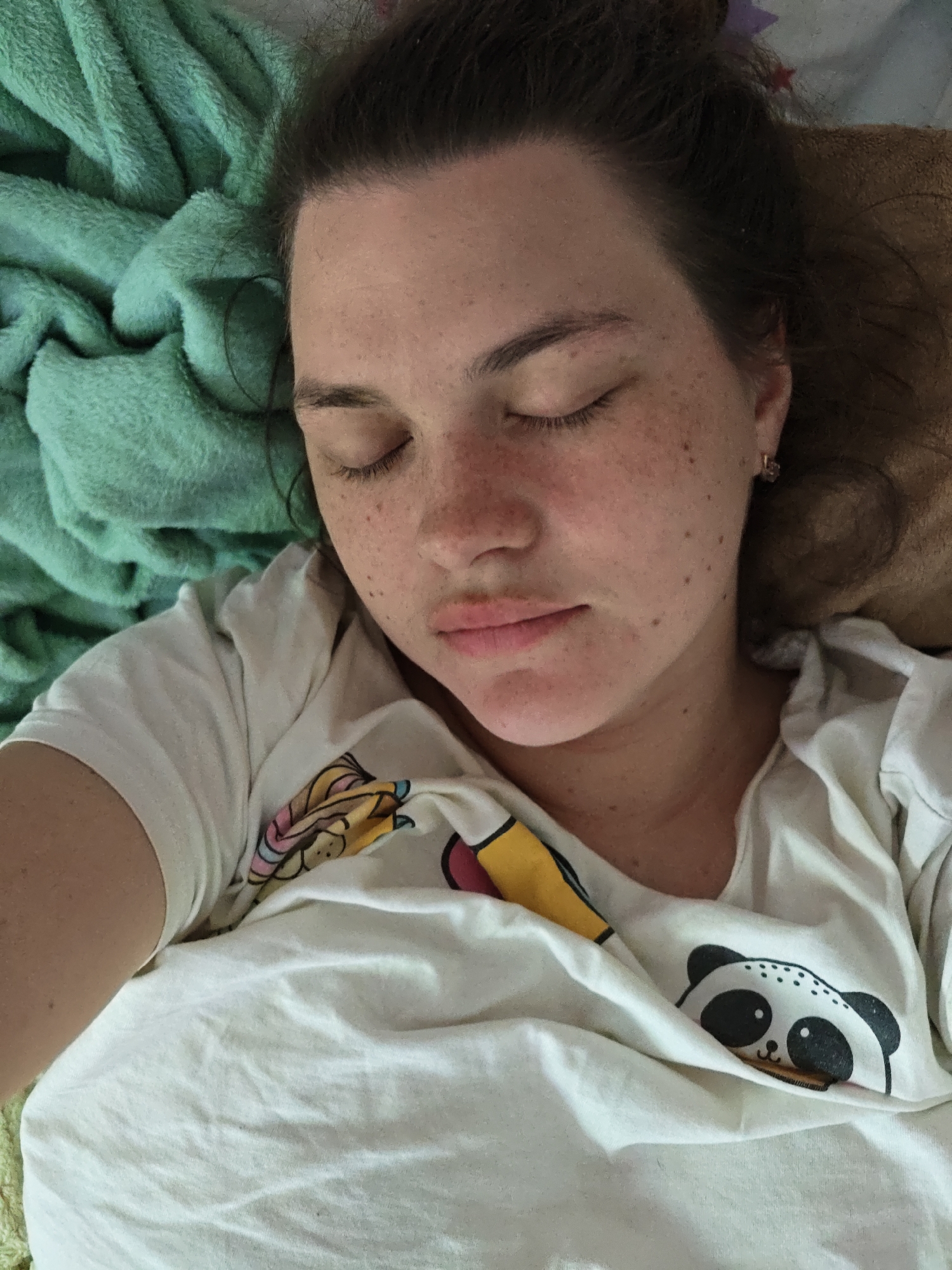 A woman is lying down with her eyes closed, seemingly sleeping. She has freckles and is wearing a white t-shirt with cartoon animal designs. She is covered by a white blanket that also has cartoon animal designs and is lying on a green textured blanket. The photo seems to have been taken at close range. The city of Chaikovsky, Russia is not mentioned in the image. The city is not apparent from the information given in the image.