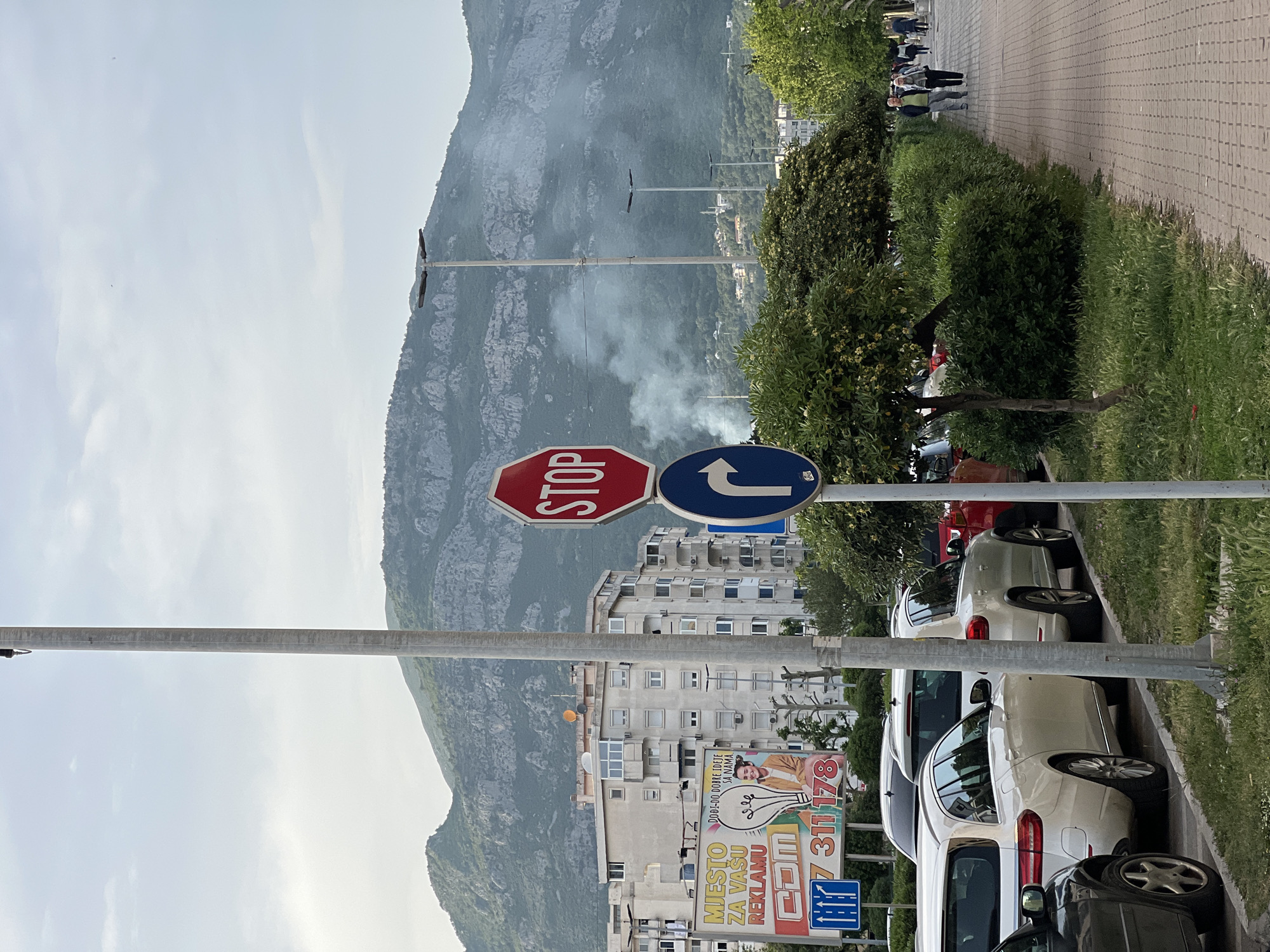 The image depicts a street scene in Bar, Montenegro, with a mountain in the background partially obscured by smoke. In the foreground, there is a stop sign and a blue right-turn-only sign. There are parked cars along the right side of the road, and a sidewalk with pedestrians. A large advertising billboard for COM is present. Above the buildings is the mountain with smoke rising from the lower part of the mountain.