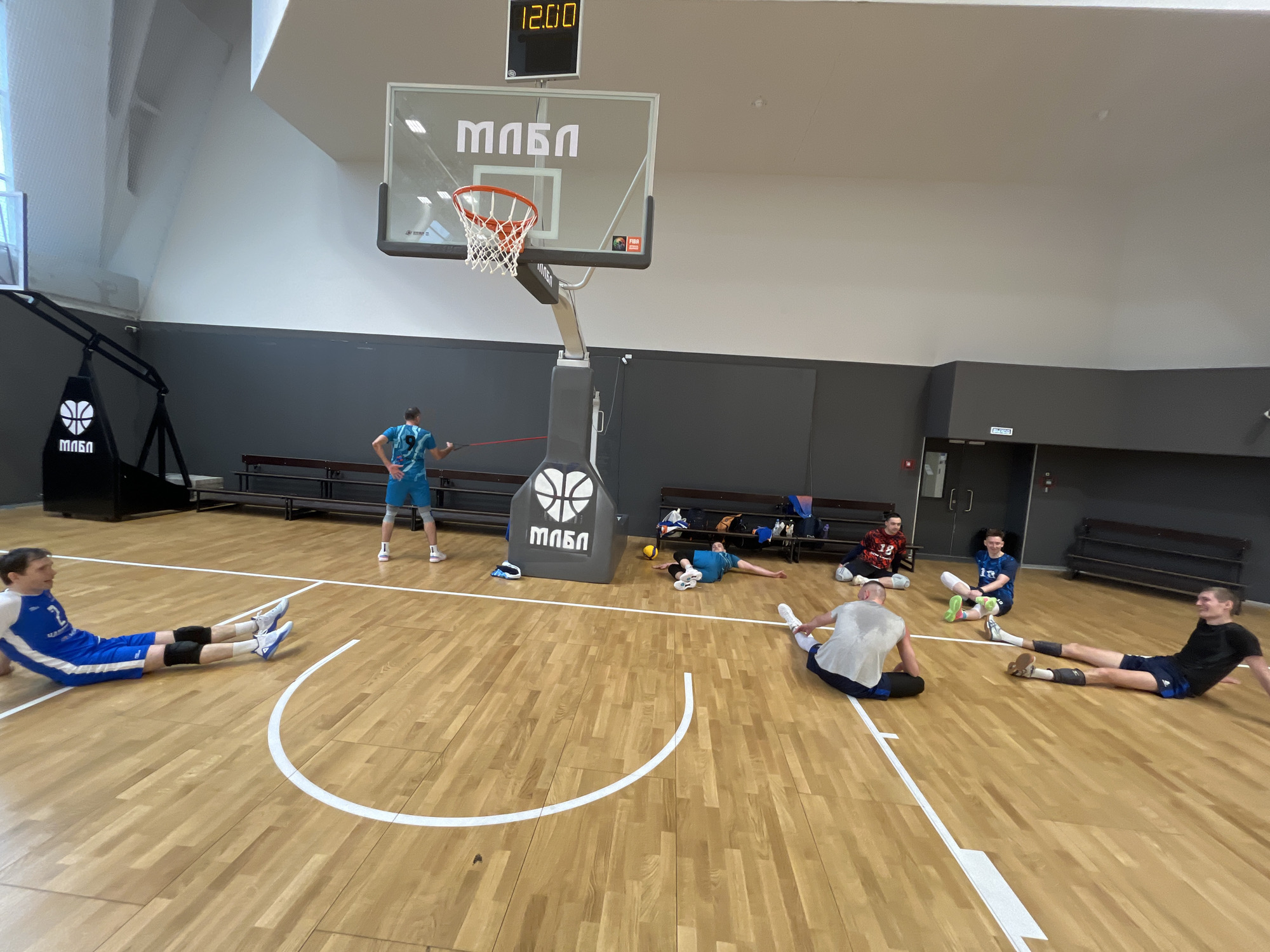 Here is a forensic description of the image:
**Location:** A gymnasium in Moscow, Russia. This is evident from the Cyrillic text "МЛБЛ" (MABL) on the basketball hoop and adjacent stand.
**Scene:** The gymnasium features a wooden basketball court marked with white lines. There are several people, mostly young men, engaged in warm-up or cool-down exercises.
**People:**
* One man is standing near the basketball hoop, using an elastic resistance band.
* Several other individuals are seated or lying on the floor, stretching. They appear to be wearing athletic attire, some with knee pads, suggesting a volleyball or basketball focus.
* Some individuals are on or near benches.
**Objects:**
* A basketball hoop with the "МЛБЛ" logo.
* A scoreboard displays the time as 12:00.
* A basketball.
* Benches line the wall in the background.
**Actions and Interactions:**
* Athletes are stretching and warming up.
* One athlete is using resistance band exercises.
* Individuals are socializing or resting in between exercises.
**Environment:**
The gymnasium is spacious and well-lit. The walls are a combination of white and dark gray. An exit sign is visible on the back wall.
