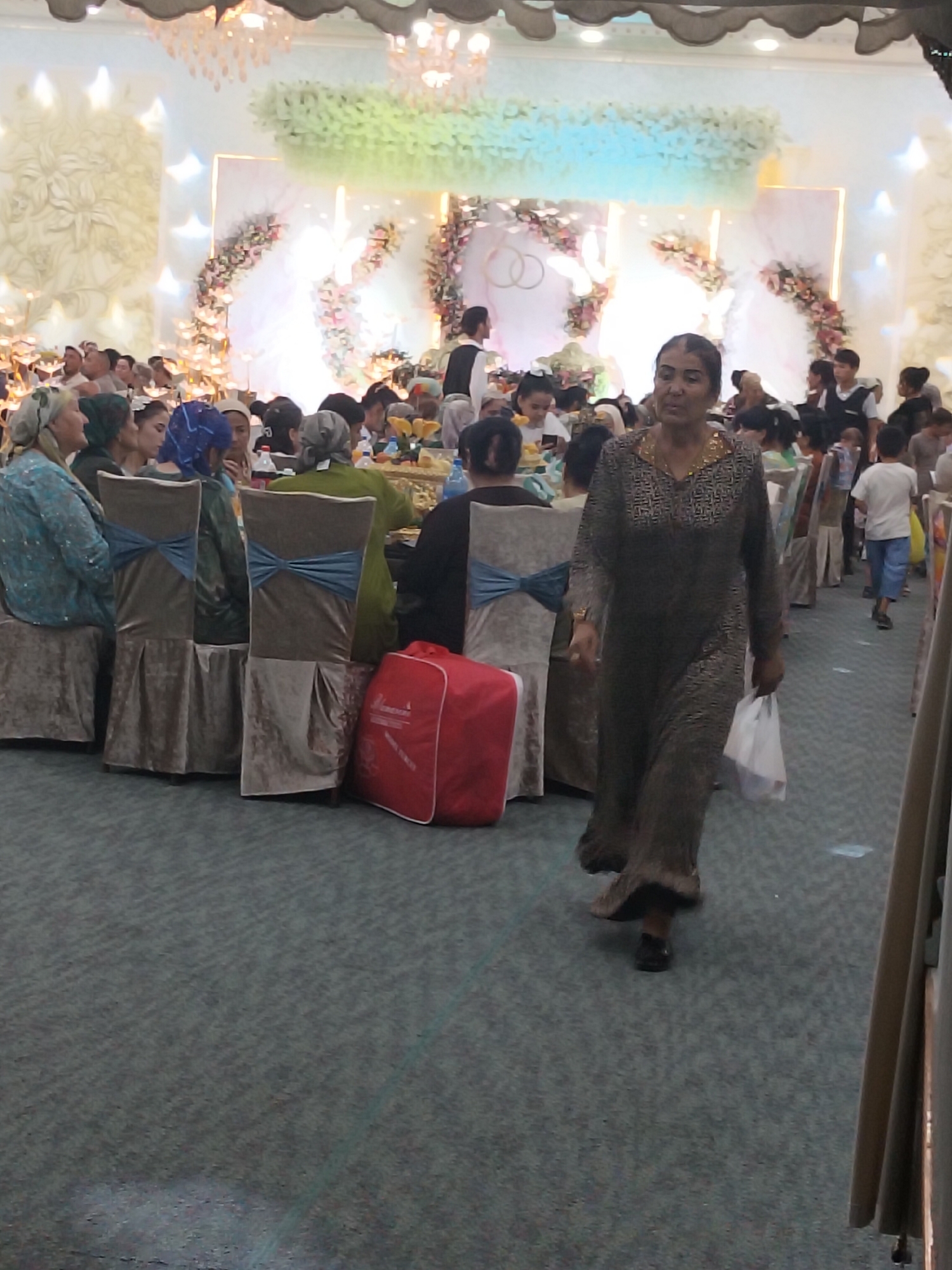 The image captures an indoor event, likely a wedding reception, taking place in Siyozi-poyon, Uzbekistan. In the foreground, a woman in a long, patterned dark dress walks from right to left, holding a white plastic bag. She is moving across a carpeted floor. Behind her, several people are seated at tables, with some chairs draped in brown velvet covers and adorned with blue bows. A large red bag is visible near the seated guests. The background features a decorated stage area with floral arrangements and illuminated panels, suggesting a formal celebration. The overall atmosphere appears festive and crowded, with numerous attendees, including children, visible in the middle ground and background.