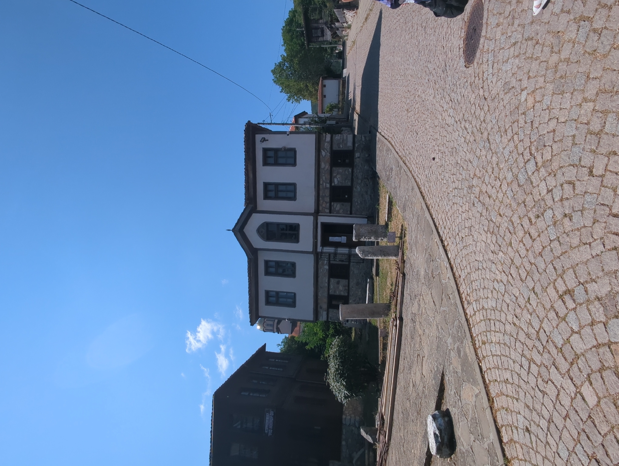 A clear, blue sky dominates the upper portion of the image. In the mid-ground, a white building with a stone base and a dark tiled roof is situated on a cobblestone street. The building has dark-framed windows on its upper level and smaller, darker windows on the lower level. To the left of this building, a dark wooden structure with a sloped roof is visible. In the foreground, a cobblestone street curves to the right, leading uphill. Scattered on the cobblestones are pieces of stone, wooden planks, and what appear to be remnants of ancient artifacts. A green bush is located to the left of the white building. In the far distance, a building with a golden dome is visible through trees. A person's legs and feet, wearing white sandals, are visible in the lower right corner of the image.