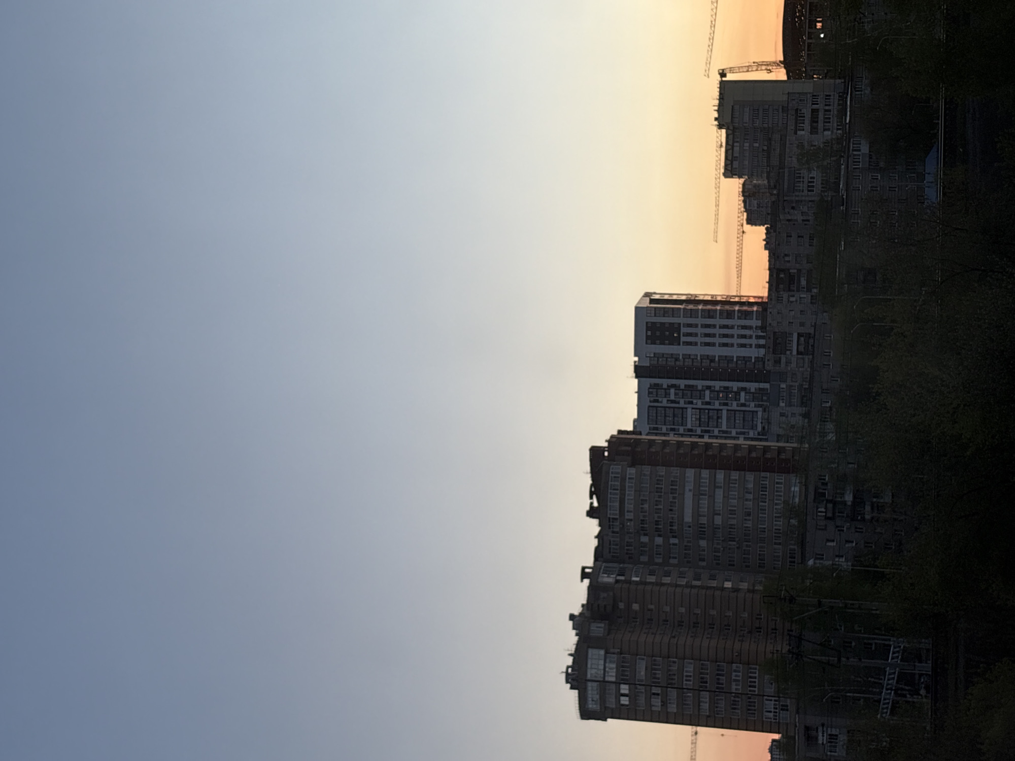 The image shows a view of a cityscape in Perm, Russia. Tall apartment buildings dominate the skyline, with construction cranes visible. The sky is a gradient of blue to orange, indicating either sunrise or sunset. Trees are silhouetted in the foreground, suggesting a view from a park or green space.