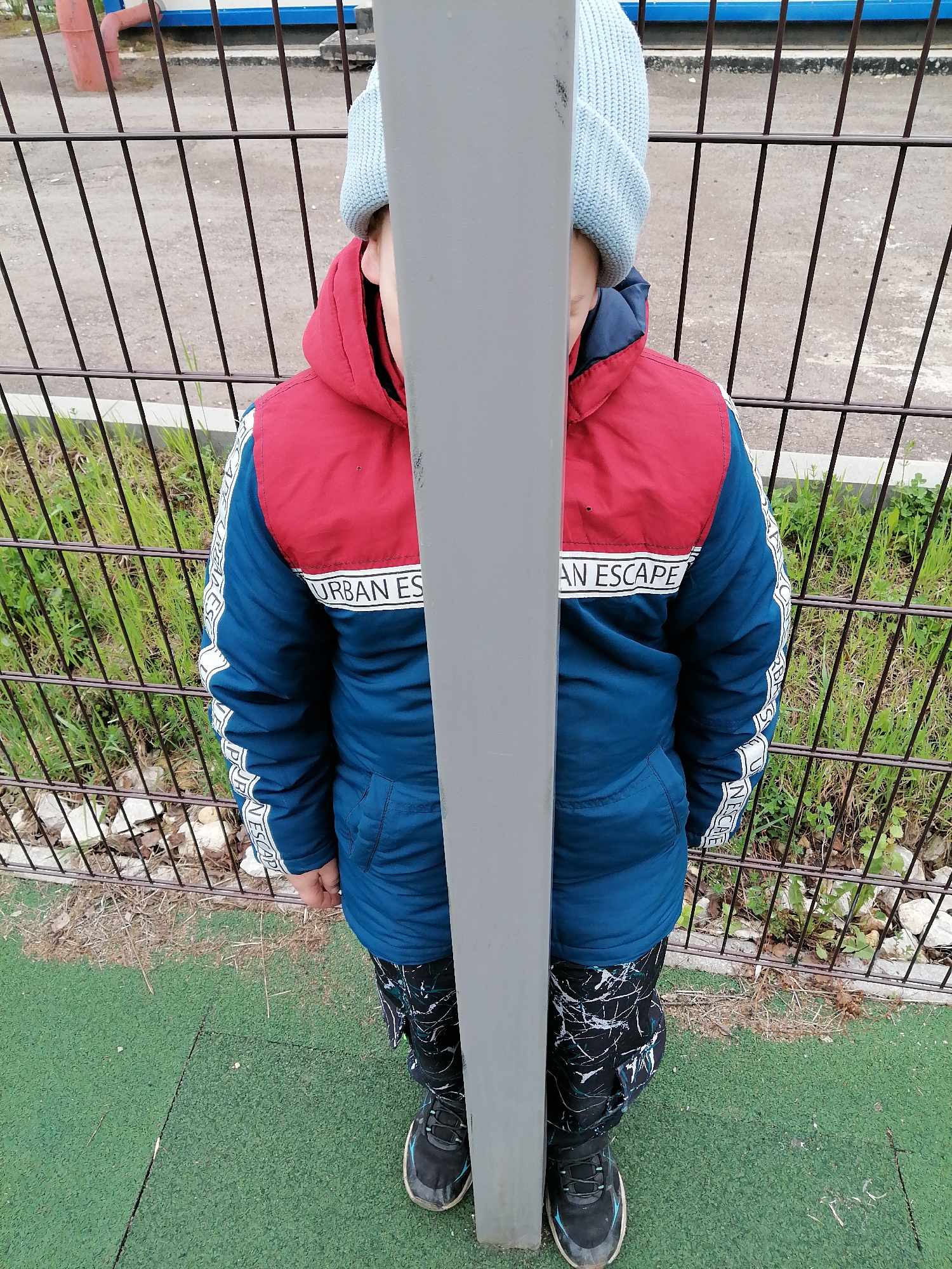 A child is standing on a green surface, possibly a playground or recreational area, in what appears to be an outdoor setting. The child is positioned behind a vertical gray metal pole, which obstructs the view of their face. The child is wearing a blue knit hat, a blue and red jacket with the words "URBAN ESCAPE" printed on a white stripe, and patterned dark pants. The child is also wearing black sneakers with light blue accents. A metal fence is visible in the background, along with grass and other vegetation. The location is Trubacheyevka, Russia.
