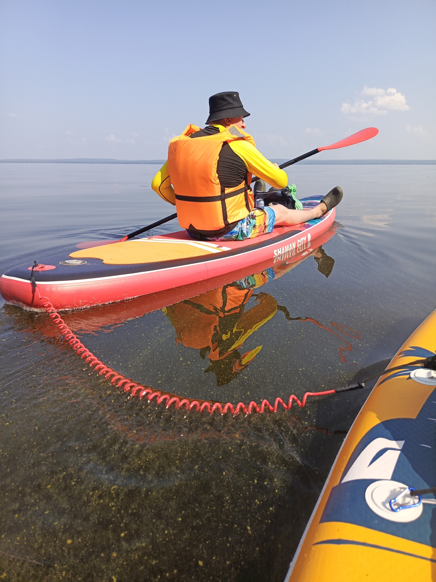 In the city of Chaikovsky, Russia, a person wearing a black bucket hat, a yellow long-sleeved shirt, and an orange life vest is seated on a red and black inflatable paddleboard. The person is holding a red paddle with a black shaft. The paddleboard has "SHAMAN CITY" written on its side. The person's legs are extended forward, and they are wearing patterned shorts and dark slip-on shoes. A red coiled leash extends from the front of the paddleboard into the water. The water is clear, revealing the sandy bottom. Another paddleboard, yellow with blue and white accents, is partially visible in the foreground on the right. The sky is clear and blue with a few white clouds. The scene appears to be on a calm body of water.