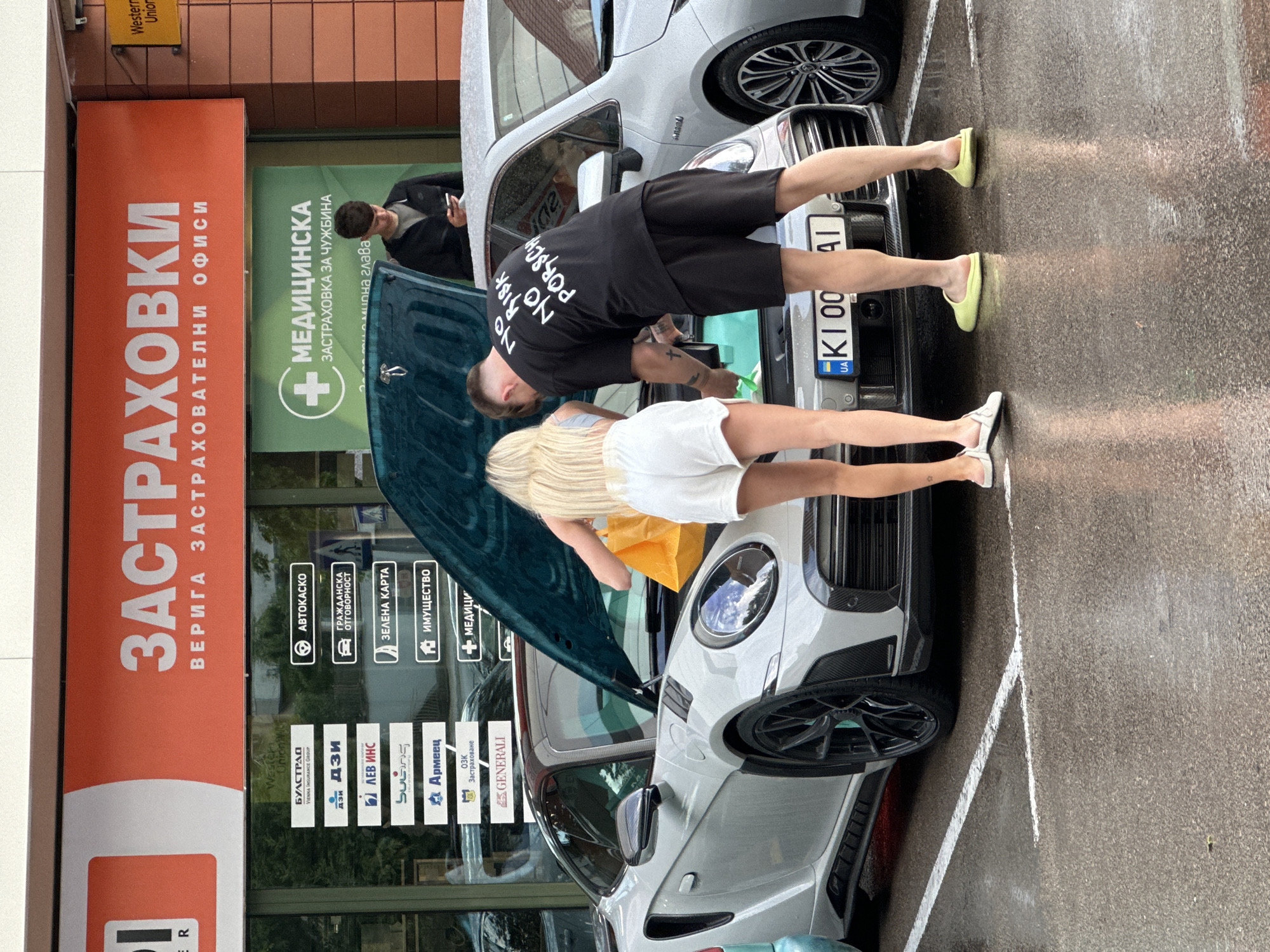 Here's a detailed description of the image:
**General Description:**
The photo is taken in an urban environment, likely in Varna, Bulgaria, given the Cyrillic script on the storefront. The focus is on two people standing in front of two vehicles with their hoods open. The weather appears wet or just after a rainfall, as the pavement is damp and reflective.
**People:**
* There are three people visible:
* A person wearing a black t-shirt with the words "NO RISK, NO PORSCH", black shorts, and yellow slip-on shoes is looking under the hood of one of the cars.
* Another person with blonde hair, wearing a white outfit and white shoes, is also looking under the hood of the same car.
* A third individual is standing near the rear of the first car, apparently looking at a smartphone or device.
**Vehicles:**
* There are two cars visible. Both appear to be a light gray or silver color.
* The front-most car has its hood open, with a dark teal/green interior visible under the hood.
* The second car is parked behind the first.
**Objects and Environment:**
* The background features a commercial building with signage in Cyrillic.
* Signs are visible for "ЗАСТРАХОВКИ" (Insurance), insurance companies, and medical insurance for foreigners "МЕДИЦИНСКА ЗАСТРАХОВКА ЗА ЧУЖБИНА".
* The pavement is marked with white parking lines.
**Actions and Interactions:**
* The two main subjects appear to be inspecting the engine compartment of one of the cars. The nature of their activity (checking, repairing, etc.) cannot be determined.
* The third person is engaged with a mobile device.
**Location:**
* Based on the signage and language, it is highly probable that the photo was taken in Varna, Bulgaria. The location is likely a commercial area where insurance services are offered.