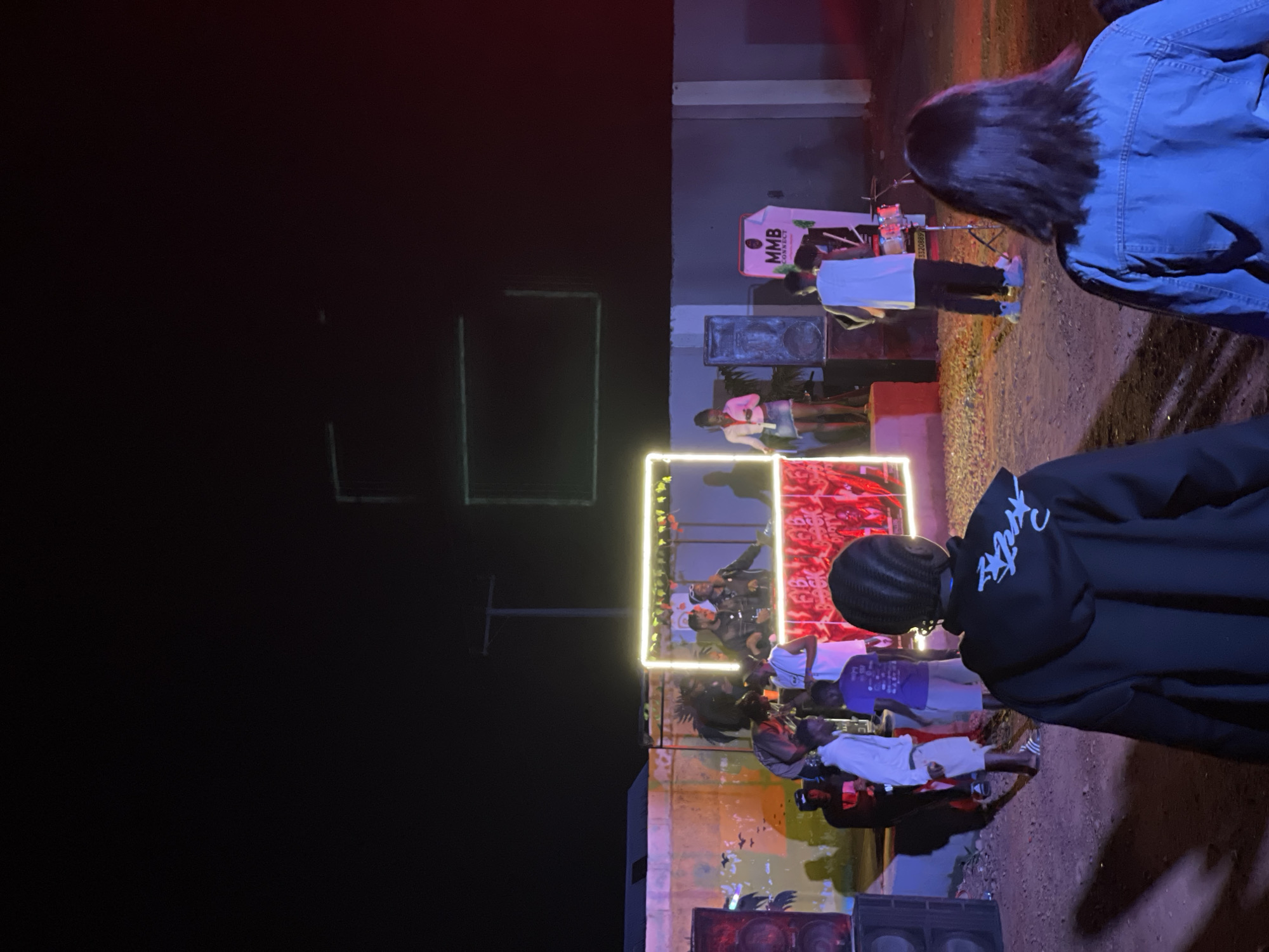 In Durumi, Nigeria, at night, a DJ set is being performed outdoors. A rectangular neon light outline illuminates the DJ booth, where several individuals are gathered. Two people are actively engaged with the DJ equipment, while others stand around. A woman in a light-colored top and denim skirt is seen near the DJ booth. To the left, a large speaker is visible, and to the right, another speaker and a sign that reads "MMB CONNECT" are present. A person is playing drums on the right side of the scene. Various individuals are scattered around the performance area, some standing and others seated. The background features a low wall and a pole with wires, suggesting an urban or semi-urban setting. The ground is covered in gravel and dirt. A dark, starless night sky forms the backdrop.