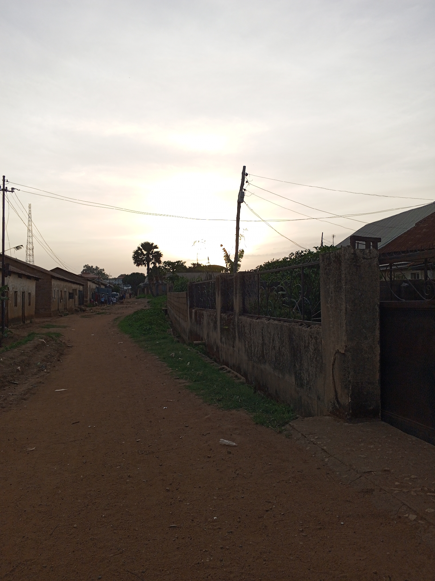 The image depicts a dirt road in Bukuru, Nigeria. The road is unpaved and bordered by buildings on the left and a concrete wall with vegetation on the right. In the distance, people are gathered. Overhead, electrical wires and a communications tower are visible against a bright sky.