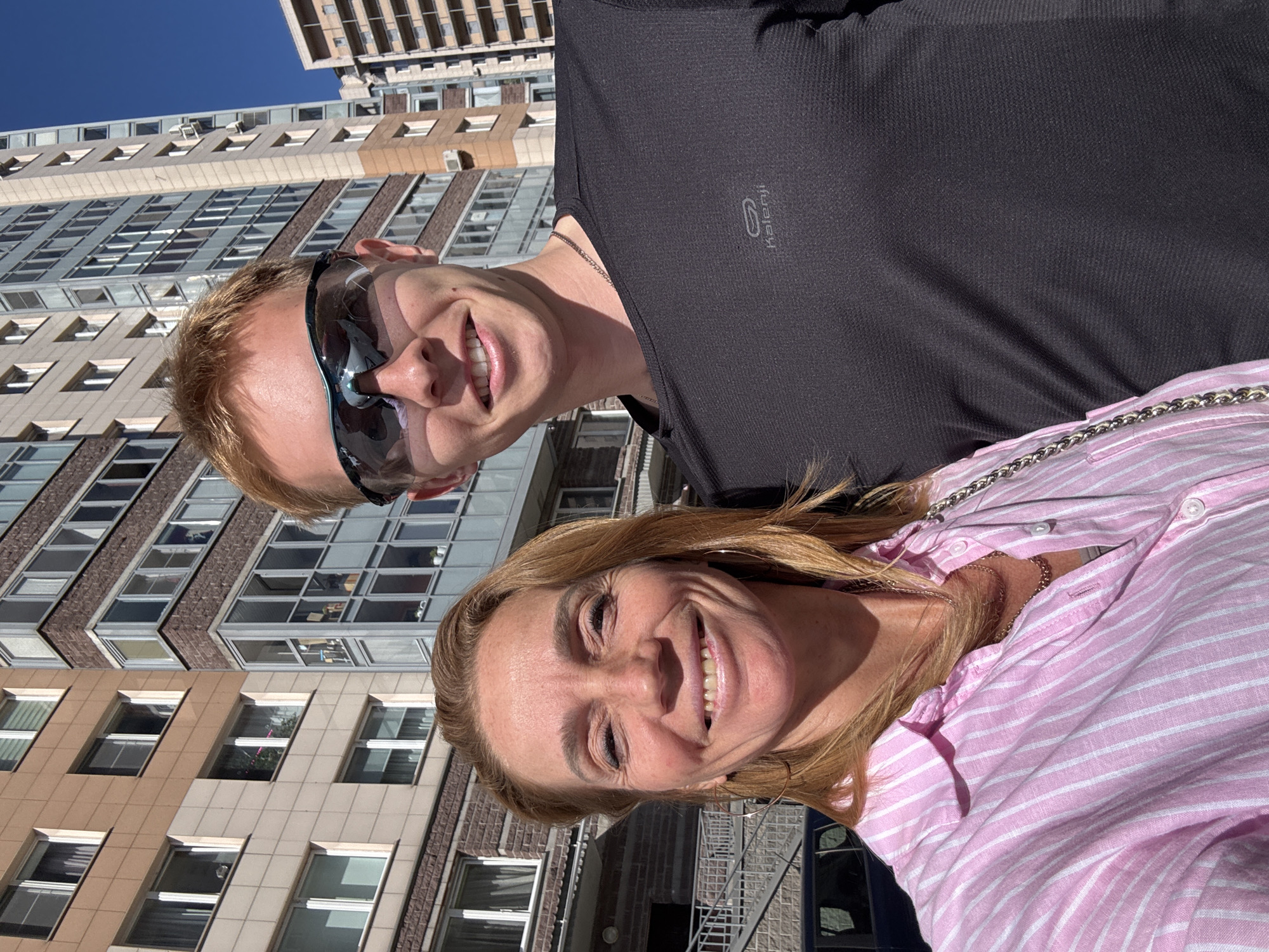 A brightly lit, vertical photo captures two smiling individuals, a man and a woman, against the backdrop of a modern urban building and a clear blue sky.
On the left, a man with light blond hair wears large, mirrored sunglasses and a black short-sleeved top featuring the "Kalenji" logo and a running figure. A thin silver chain is visible around his neck. He is smiling broadly towards the camera.
To the right and slightly in front, a woman with long, light brown hair also smiles warmly. She is wearing a pink and white striped button-down shirt, with a gold chain visible around her neck and a silver-chained bag strap resting on her shoulder.
The background consists of a tall, multi-story building characterized by rows of rectangular windows and alternating sections of beige/light brown and darker brown paneling, resembling brickwork. Several air conditioning units are mounted below some windows. Above the building, a pristine blue sky is visible, indicating bright, sunny weather during daytime. Both individuals appear happy and cheerful.