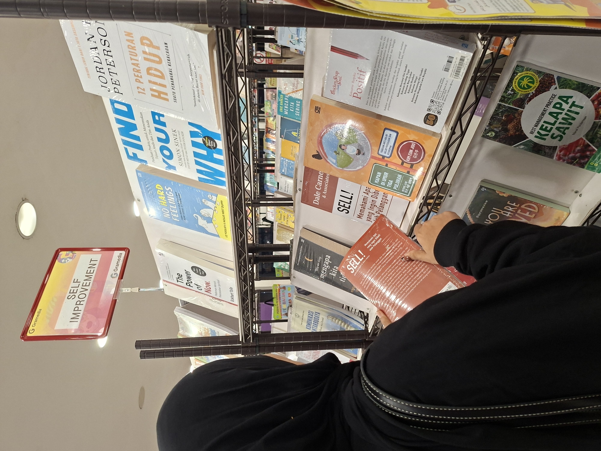 A person wearing a black hijab is looking at books on a shelf in a bookstore in Lhokseumawe, Indonesia. The sign above the books says "SELF IMPROVEMENT". The titles of the books visible on the shelf include: "12 PERATURAN HIDUP" by Jordan Peterson, "FIND YOUR WHY" by Simon Sinek, "The Power of Now" by Eckhart Tolle, "Mengapa Kita Sering" by Eric Barker, "SELL!" by Dale Carnegie & Associates, "Chicken Soup for the Soul: Kekuatan Berpikir Positif", and "BEST MANAGEMENT PRACTICE KELAPA SAWIT". The bookstore is well-lit and the shelves are organized. The person is holding an orange book that has the title "SELL!".