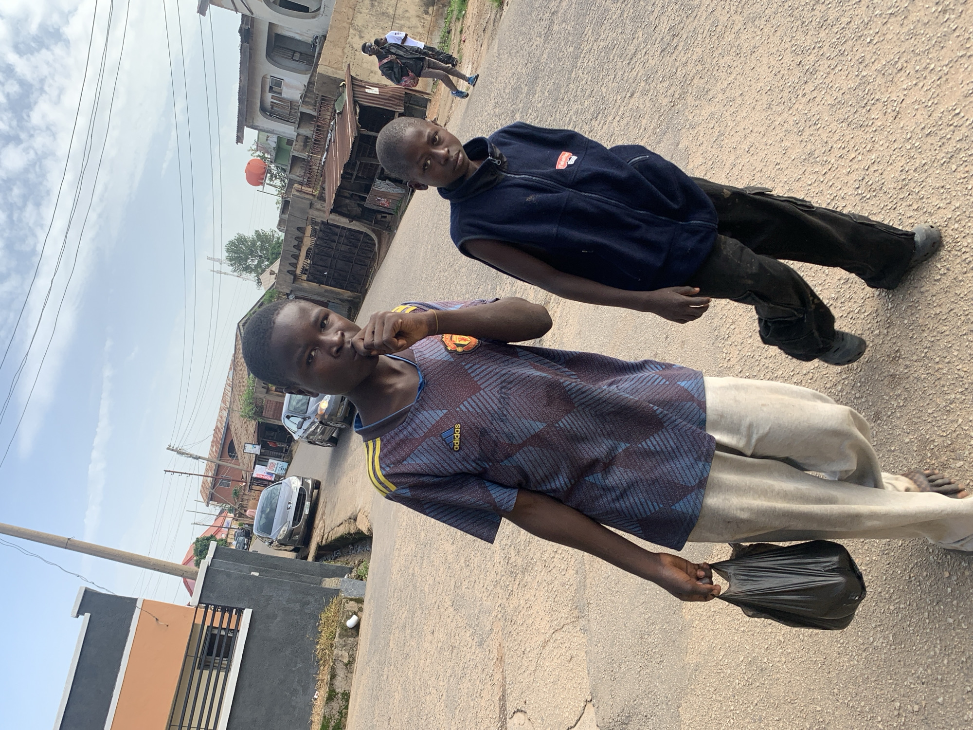Two young boys are walking on a paved street in Jos, Nigeria. The boy on the left, in the foreground, wears a blue and maroon Adidas shirt and light grey pants. He is carrying a black plastic bag in his left hand and has his right hand near his mouth. The boy on the right wears a dark blue vest over a black shirt and dark pants. He is looking towards the camera. In the background, there are several buildings, including a two-story structure on the left and smaller, single-story buildings with corrugated metal roofs further down the street. A few cars are parked or driving on the road. The sky is partly cloudy. The overall scene depicts a typical street in a Nigerian city.