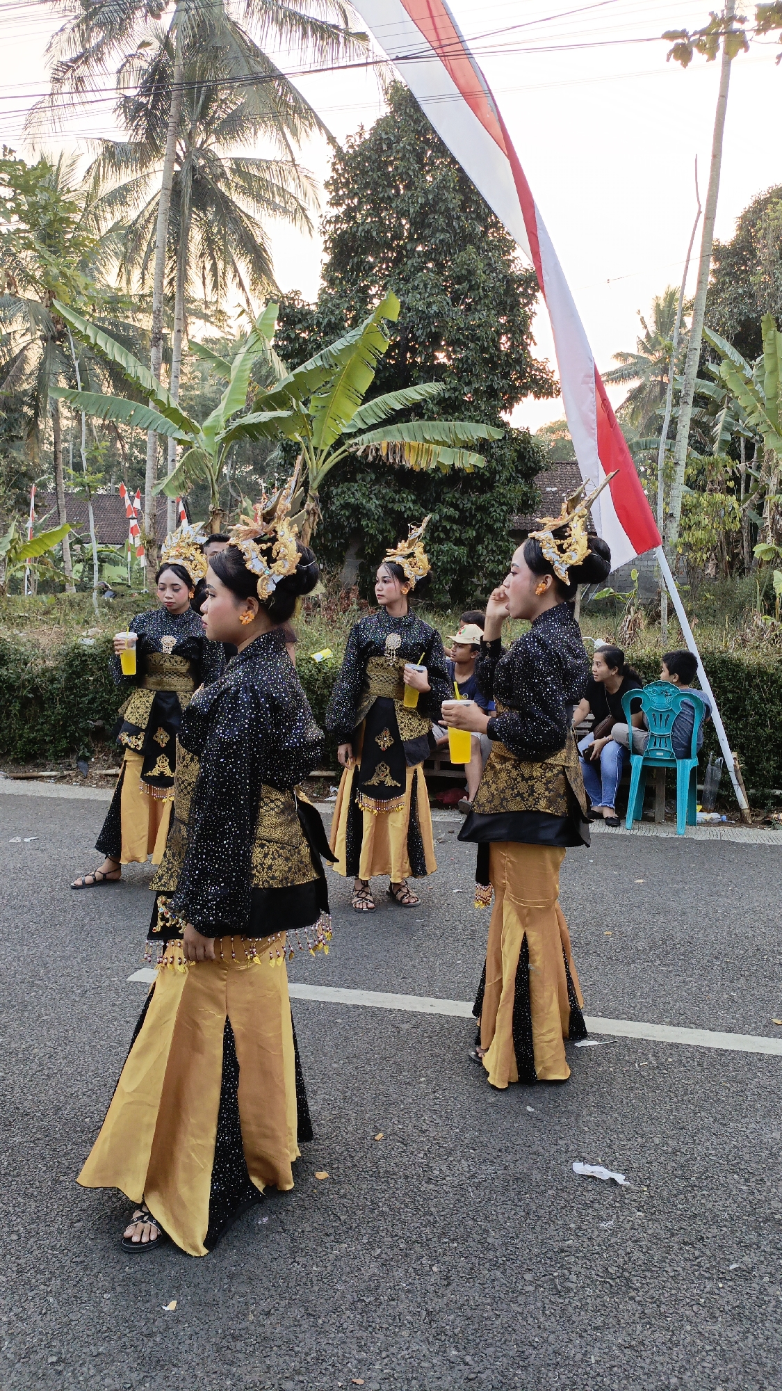 In Krajan Lor, Indonesia, five young women in elaborate traditional costumes stand on an asphalt road. Their costumes feature black tops adorned with gold embellishments and bejeweled headpieces. The women are also wearing long, flowing gold skirts with black underskirts. Two of the women are holding yellow cups. In the background, palm trees and other lush vegetation line the roadside. A red and white Indonesian flag is prominently displayed, partially obscuring the sky. Several onlookers, including children, are seated on benches and a blue plastic chair in the distance. The scene appears to be part of a local celebration or cultural event.