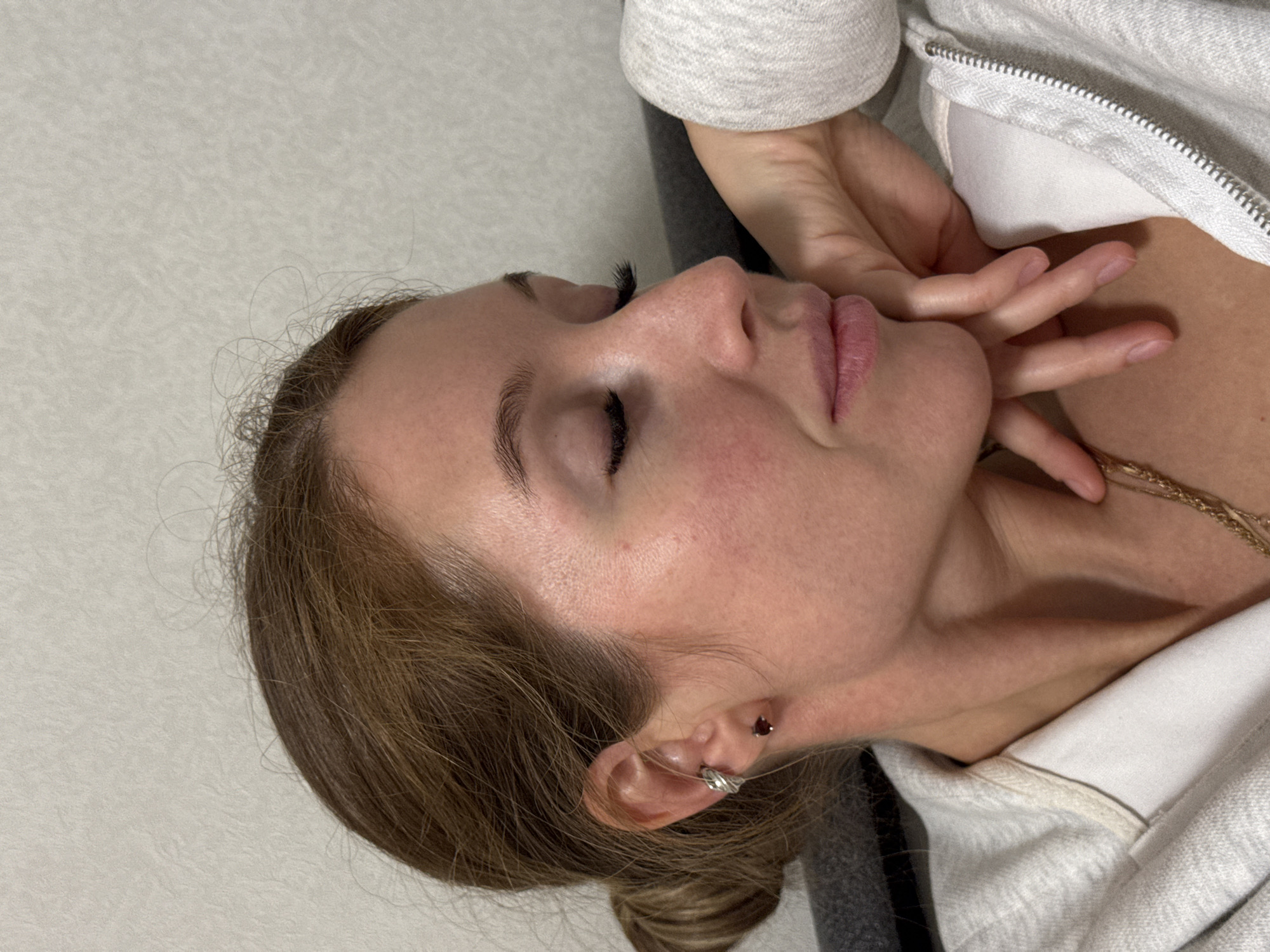 This is a close-up, profile portrait of a person, likely female, shown resting in an indoor setting in Chaikovsky, Russia. The individual's head is tilted, and their eyes are closed, suggesting a moment of rest, sleep, or contemplation. Their right hand is gently placed against their chin and cheek.
The person has brown hair, which appears to be styled away from the face, with some strands visible near the forehead and ear. Notable facial features include long, dark eyelashes, defined eyebrows, a small nose, and full, pink-toned lips that are slightly parted. The skin on the cheek and around the nose shows a hint of redness.
The right ear is adorned with two piercings: a small, dark heart-shaped stud in the upper helix and a silver-toned cuff or ring on the mid-helix. Around the neck, a delicate gold-colored chain necklace is visible. The person is wearing a light gray or off-white hooded sweatshirt with a zipper detail, layered over a white garment.
The background consists of a plain, light-colored, subtly textured wall or surface. A darker, possibly gray, surface is visible beneath the person's shoulder area, likely a chair or bed. The overall mood conveyed is calm and serene. No text is visible in the image.