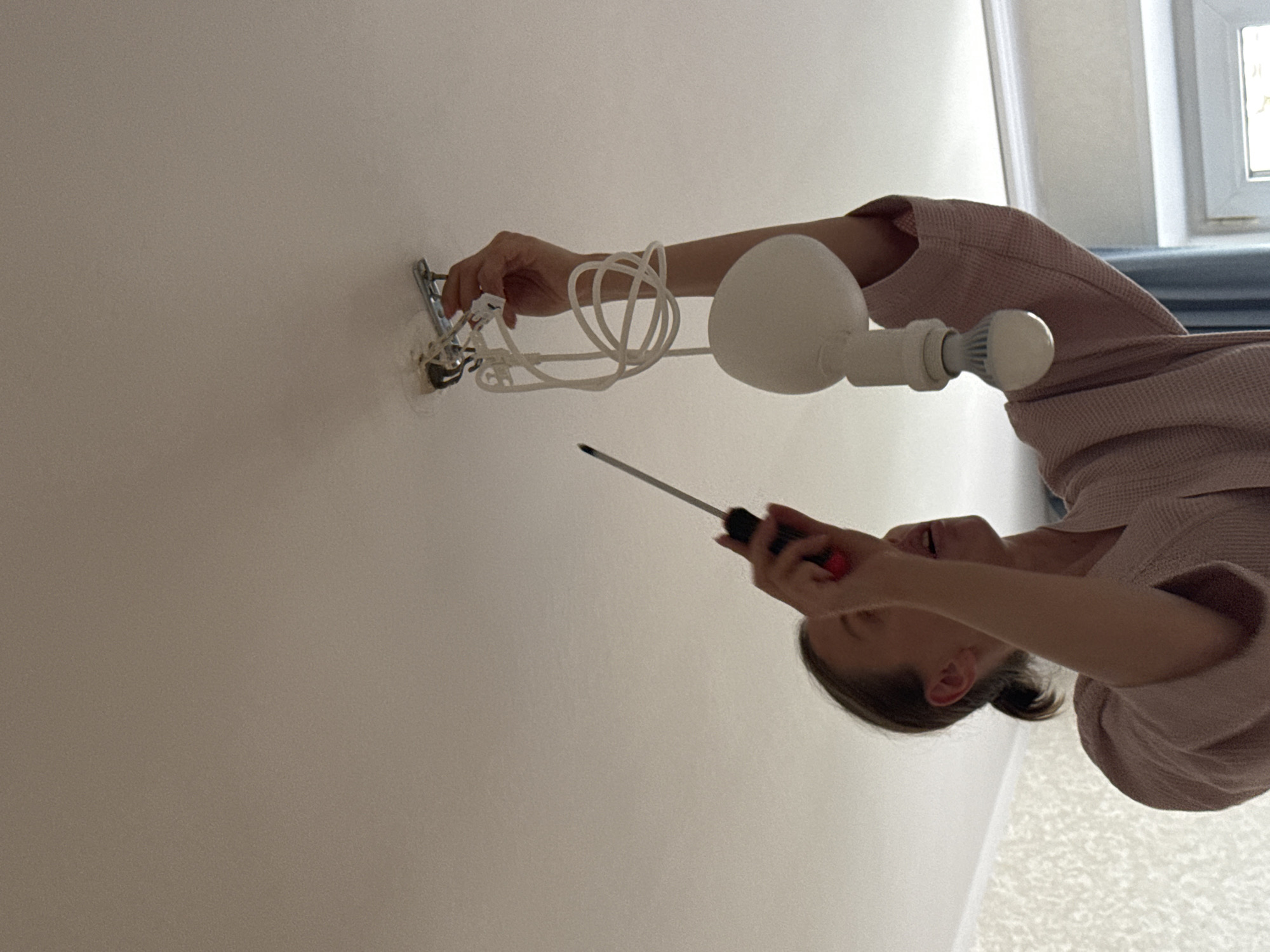 In Moscow, Russia, a woman is pictured from below, on the ceiling of an interior room, installing a new light bulb. She is wearing a pink robe and her hair is tied back in a ponytail. She holds a screwdriver in her right hand and is in the process of screwing in a white LED light bulb, which is attached to a white cord and hanging from a metallic fixture mounted on the ceiling. The room's ceiling is white and appears to be made of stretch fabric. The background shows a textured wallpaper on the left and a window with blue curtains on the right.