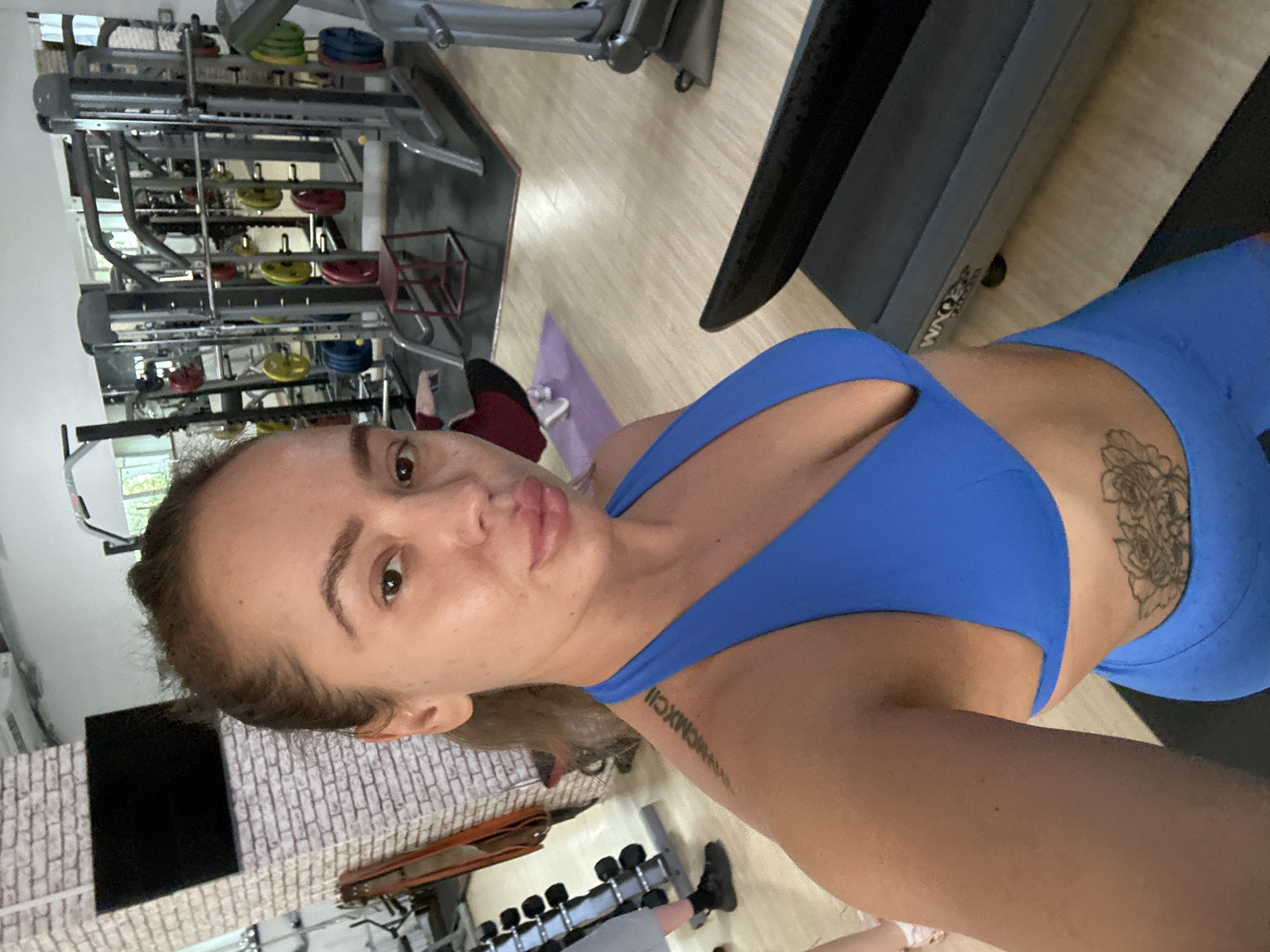 A woman is taking a selfie in a gym. She is wearing a blue sports bra and blue leggings. She has a tattoo on her left shoulder and a floral tattoo on her abdomen. In the background, there is gym equipment, including weight machines and dumbbells. There are also other people in the gym, though they are out of focus. The gym appears to be well-lit and spacious. The woman is looking directly at the camera with her lips slightly parted.