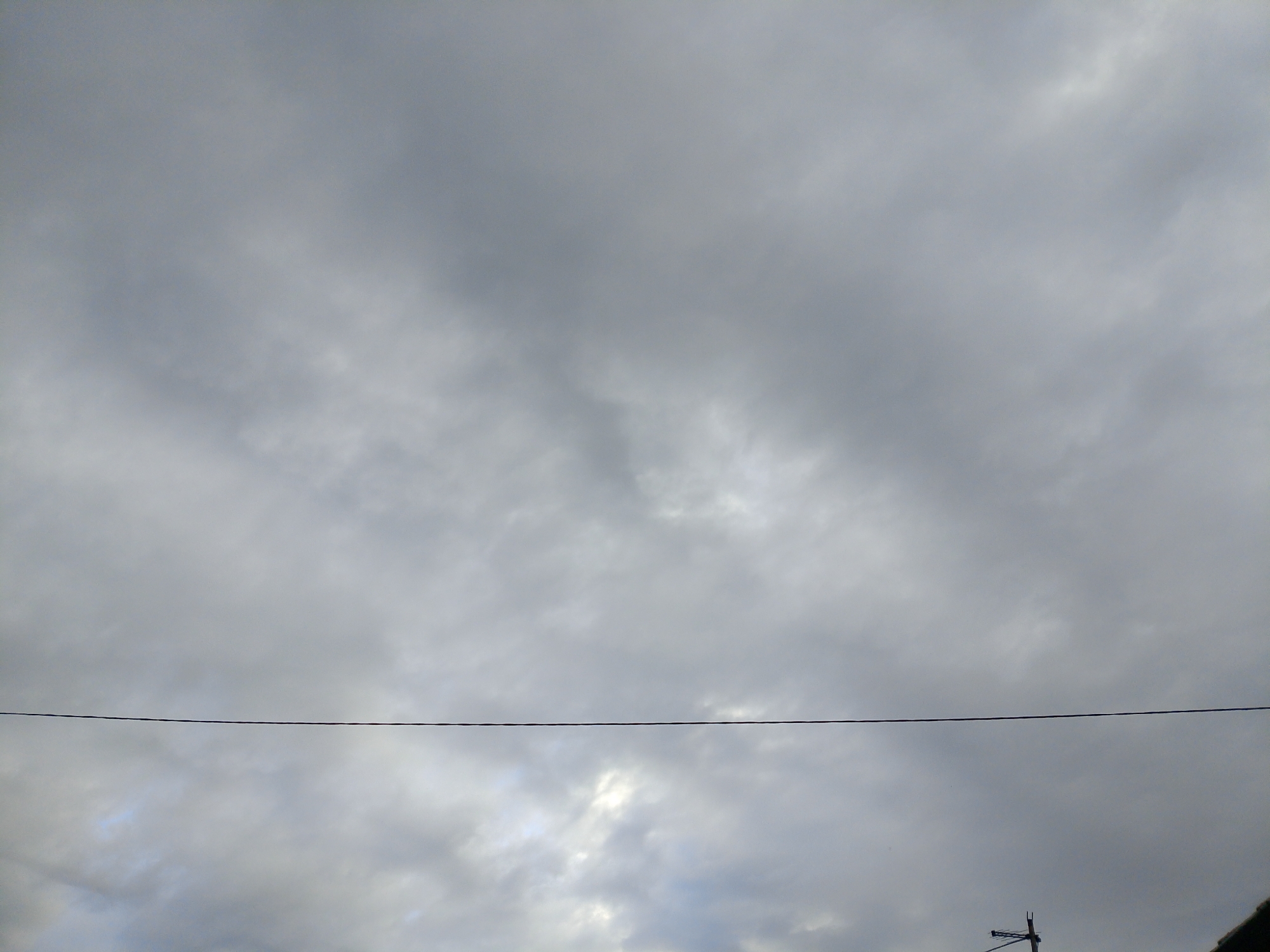 The image shows a cloudy sky with a thin horizontal wire stretching across the frame. In the lower right corner, a utility pole with what appears to be an antenna is partially visible, along with the edge of a dark roof. The overall scene suggests an outdoor view from a potentially urban or suburban setting. The location in Закалтус, Russia, is not visually discernible from the provided image.