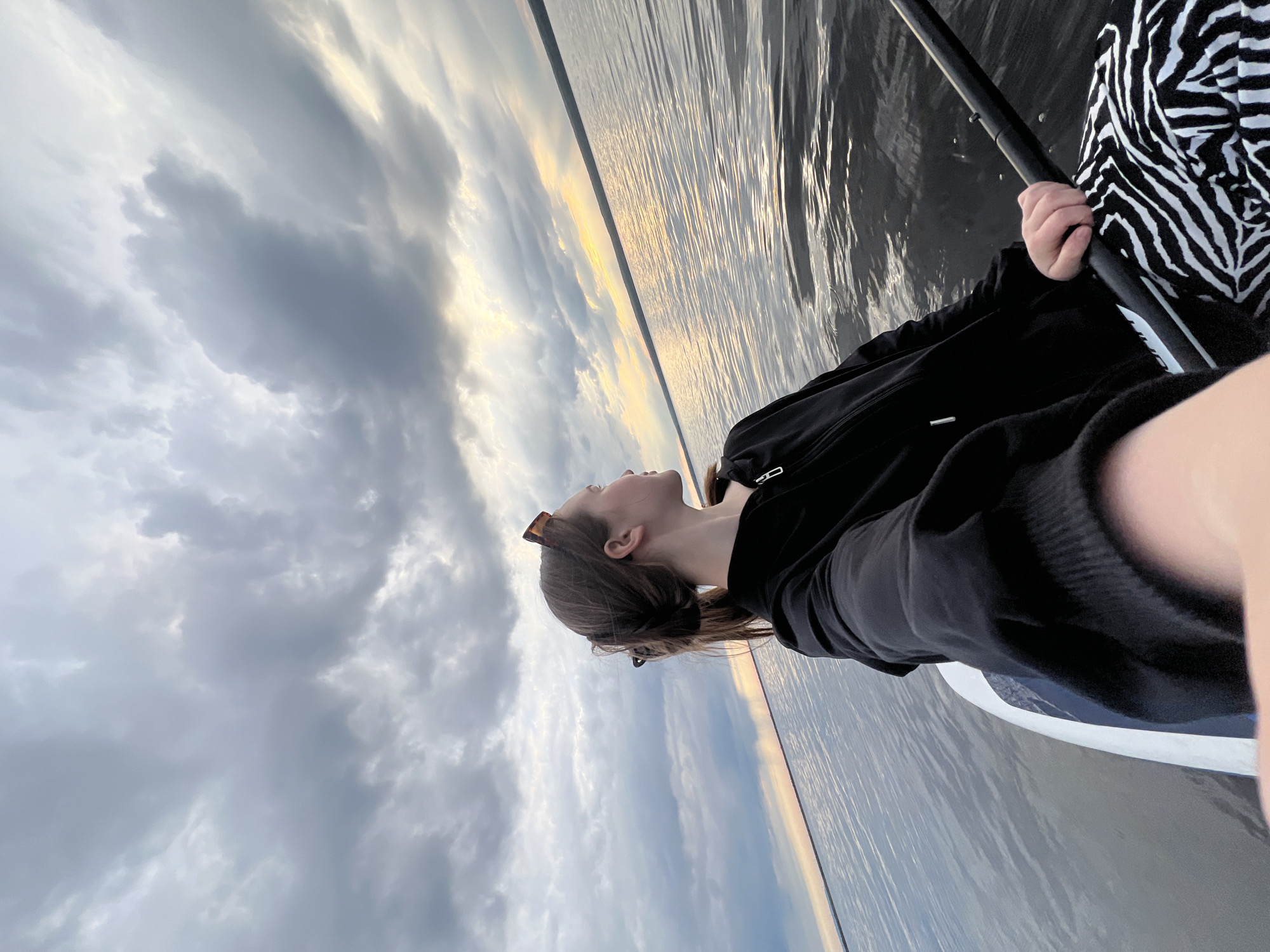 The image captures a person paddleboarding on a vast body of water under a dramatic sky. The main subject, seen in profile, is a person with dark hair, wearing a black hooded top over a white and black zebra-print garment. Sunglasses are perched on their head. They are holding a black paddle with their right hand and are looking upwards and towards the distant horizon.
The setting is a wide, calm expanse of water that reflects the cloudy sky. The horizon line features a strip of golden-orange light, suggesting either sunrise or sunset. Above the horizon, the sky is filled with voluminous, grey, overcast clouds, interspersed with brighter patches. The person is positioned on a white paddleboard, the edge of which is visible in the foreground.
The activity taking place is paddleboarding or boating, with the individual seemingly at rest, observing the serene and picturesque natural environment. The time of day is twilight, characterized by the soft, diffused light and the striking colors along the horizon. The location context of Chaikovsky, Russia, suggests this scene is likely on a large lake or wide river. There is no visible text in the image. The overall mood is one of peaceful contemplation amidst nature.