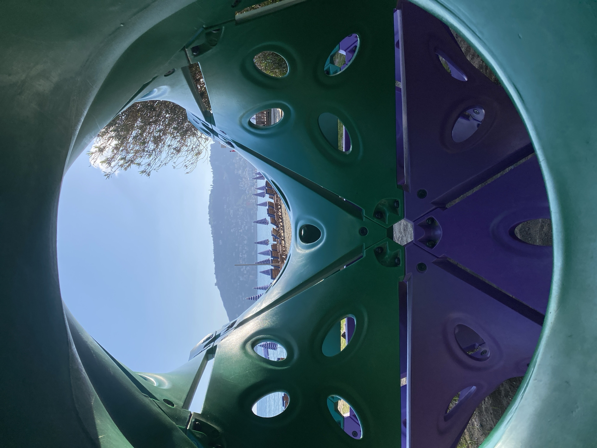 The image captures a vibrant view from inside a brightly colored playground tunnel or climbing structure, looking out towards a scenic coastal landscape.
In the immediate foreground, the interior walls of the playground equipment are visible. The left side is a solid green, while the right side, further within the structure, is a contrasting purple. Both the green and purple panels feature multiple oval and circular cutouts, some with raised edges, allowing light and glimpses of the outside to pass through. Hexagonal bolts are visible where different panels of the structure connect.
Through these openings and from the general perspective, a clear, sunny day unfolds in the background. A bright blue sky dominates the upper-left portion of the view. Below and towards the right, a sandy beach is visible, adorned with neat rows of blue and white striped beach umbrellas and accompanying sun loungers. Beyond the beach, a body of water, likely the sea, stretches out, and in the distance, a range of green hills or mountains rises, partially obscured by a slight atmospheric haze.
The scene depicts no specific activity or people, but the presence of the playground and beach setup suggests a leisure or recreational environment. The bright, clear lighting indicates it is daytime with good weather. The combination of beach, sea, mountains, and resort-style umbrellas strongly points to a coastal vacation destination, consistent with Alanya, Turkey. There is no visible text in the image.