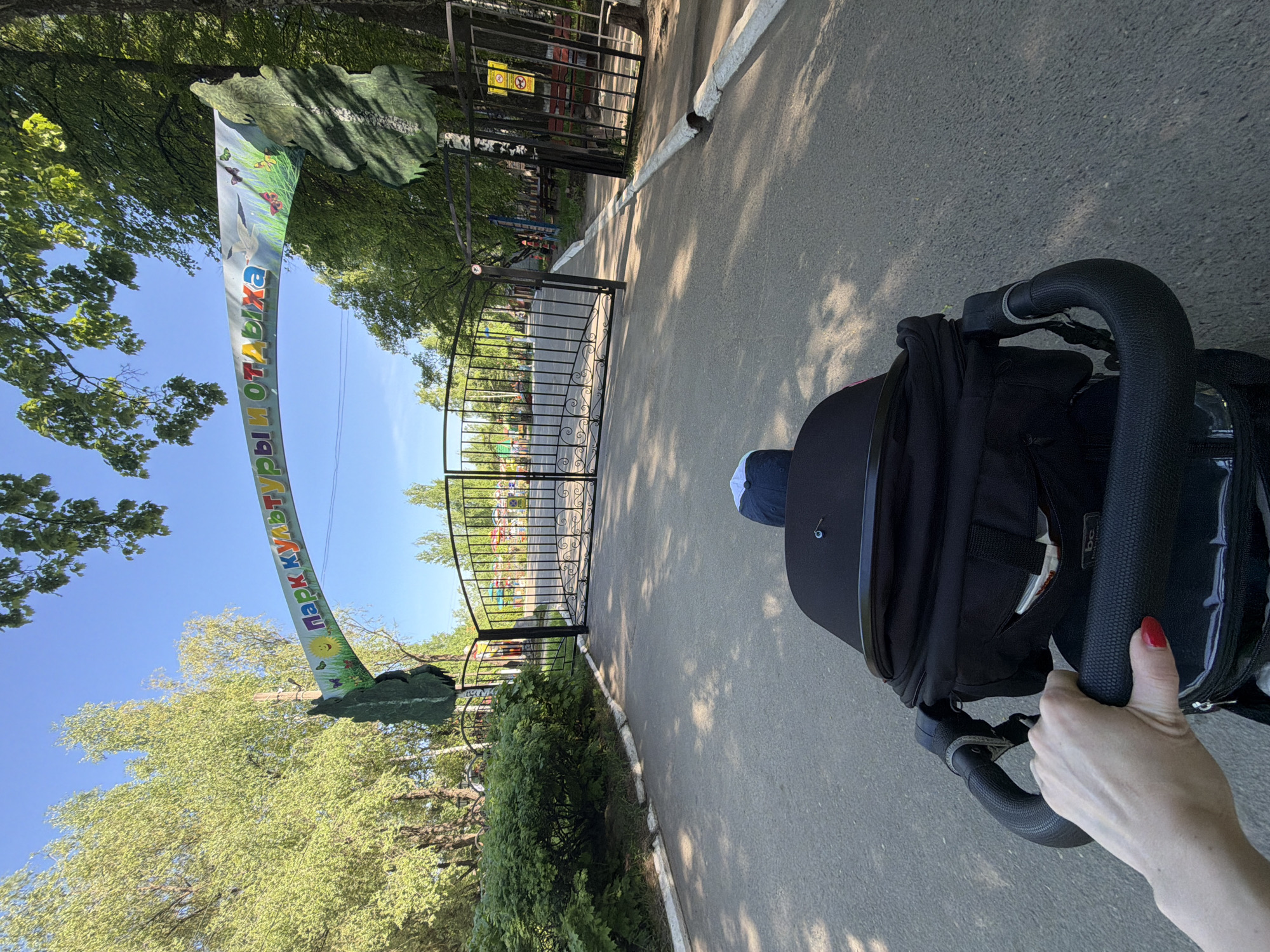 A person pushes a baby stroller toward the entrance of a park. The gate and a decorative archway with the Cyrillic inscription "Парк культуры и отдыха" (Park of Culture and Rest) mark the entrance. The park is likely located in the city of Chaikovsky, Russia, as the inscription is in Russian. The stroller is black, and a small child is visible inside, wearing a blue cap. The person pushing the stroller has a hand visible with a red manicure. The surroundings include trees and pavement.