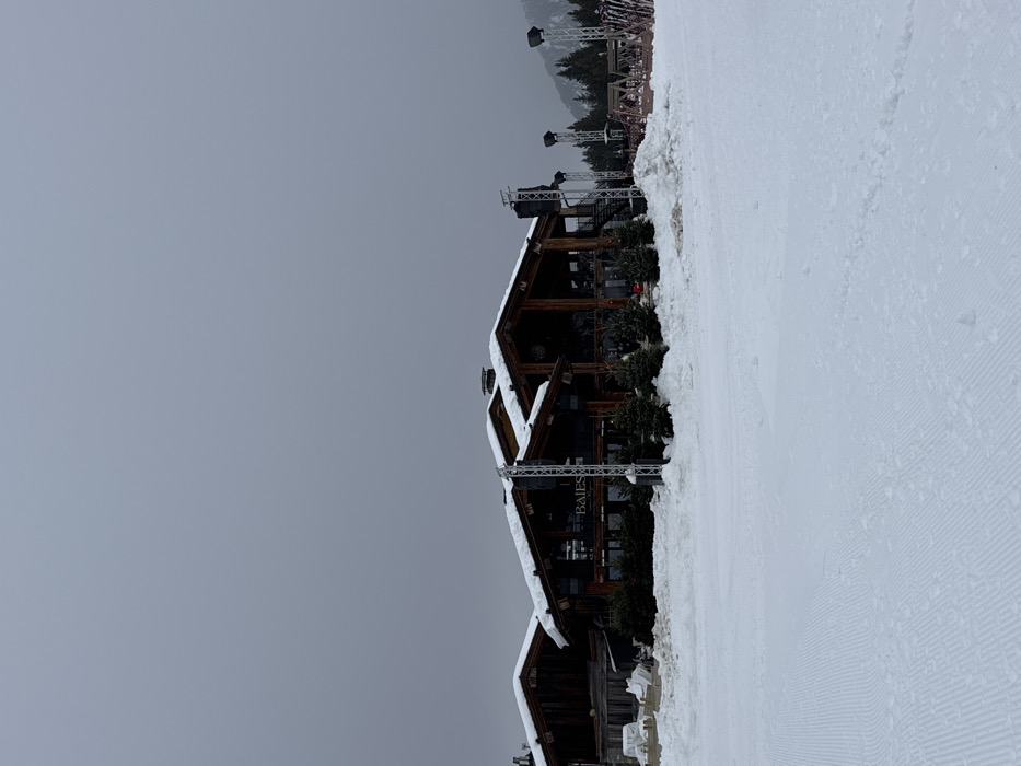 The image displays a wide-angle view of a snow-covered landscape at a ski resort in Courchevel, France, under an overcast sky. In the midground, a large, multi-section chalet-style building with dark wooden siding and steeply pitched, snow-covered roofs dominates the scene. The building features numerous large windows and a visible sign on its facade that reads "BAIES." Several small evergreen trees are planted directly in front of the building.
Flanking the building are multiple tall, metallic truss structures, some equipped with black loudspeakers and lighting fixtures, indicating an outdoor event setup. To the immediate right of the building, a substantial pile of snow is accumulated, and further right, stacked ski or snowboard racks are partially visible.
The foreground consists of an expansive, groomed snowfield, exhibiting distinct parallel ridges from snow-grooming equipment, characteristic of a ski slope. Scattered small clumps of snow are present across this groomed surface.
In the background, a dense forest of coniferous trees covers a gentle slope, beyond which faint outlines of higher, rugged mountains are visible, partially obscured by the heavy, uniformly grey overcast sky. No individuals are present, and no actions or interactions are depicted in the scene.
