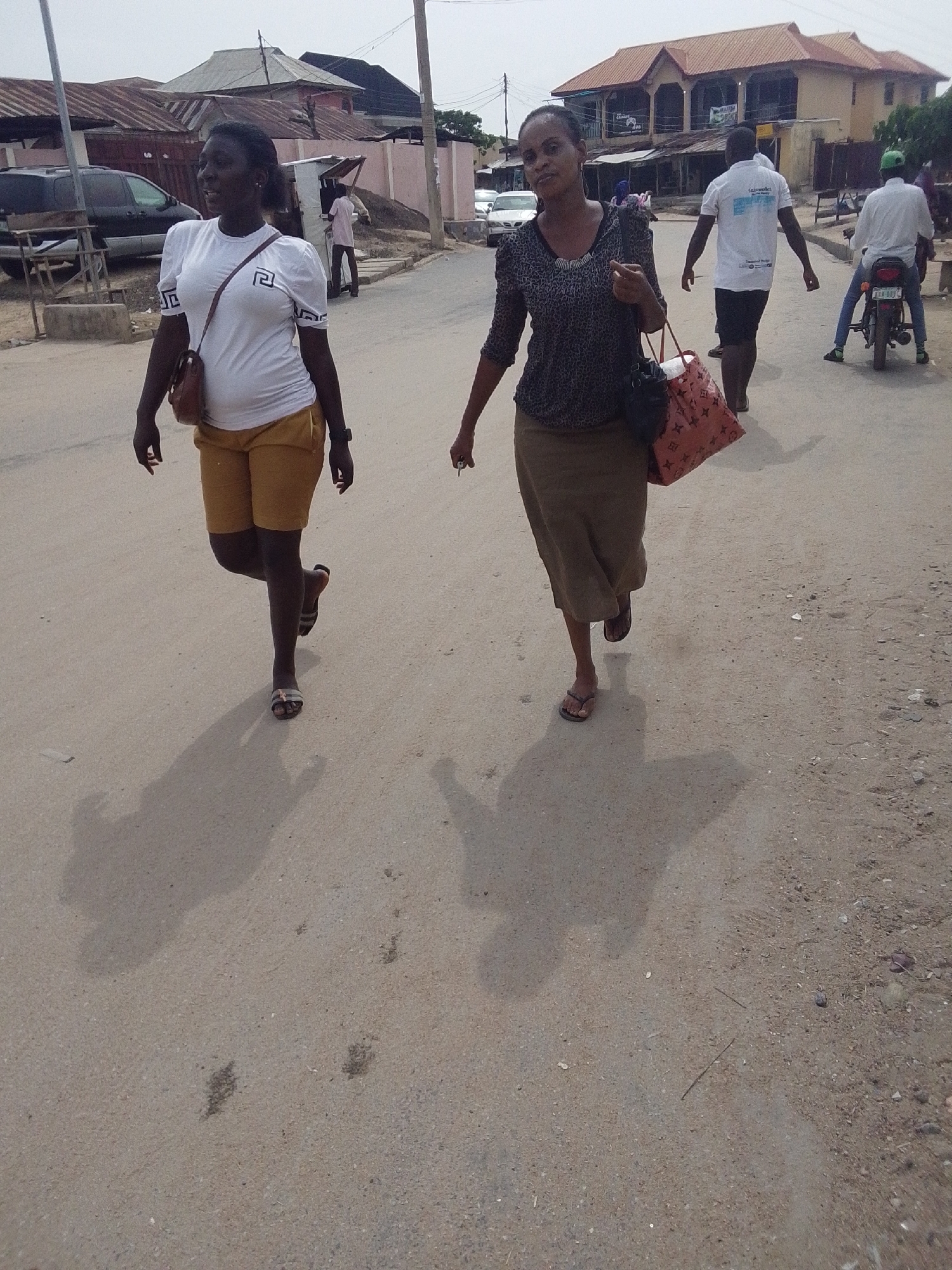 Two women are walking on a street in Lokoja, Nigeria. The woman on the left wears a white t-shirt, yellow shorts, and sandals, and carries a shoulder bag. The woman on the right wears a patterned blouse, a long skirt, and sandals, and carries two bags. Several people, a car, and a motorcycle are visible in the background. There are various buildings on either side of the street. The road is unpaved.
