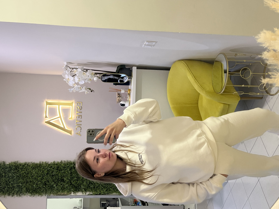 A female subject is photographed taking a mirror selfie within an interior space identified as an 'ESTETICA' clinic or salon, located in Unknown, Unknown. The subject, with light brown hair, is wearing a cream-colored hooded sweatshirt and matching sweatpants. She holds a silver-colored smartphone in her left hand, pointed towards the mirror, with her right hand in her pocket. The background features a pale-colored wall adorned with a backlit gold 'EA ESTETICA' sign and a vertical panel of artificial green foliage. A bright yellow ribbed velvet armchair and a small gold-framed side table with a mirrored top are visible. A white shelf behind the subject holds a white artificial orchid arrangement, a gold decorative bowl, a reed diffuser, various small bottles, and a white dome security camera. White tiled flooring is visible, and the lower right foreground shows fluffy dried grass decor. A coffee machine and white shelving are partially visible on the far left.