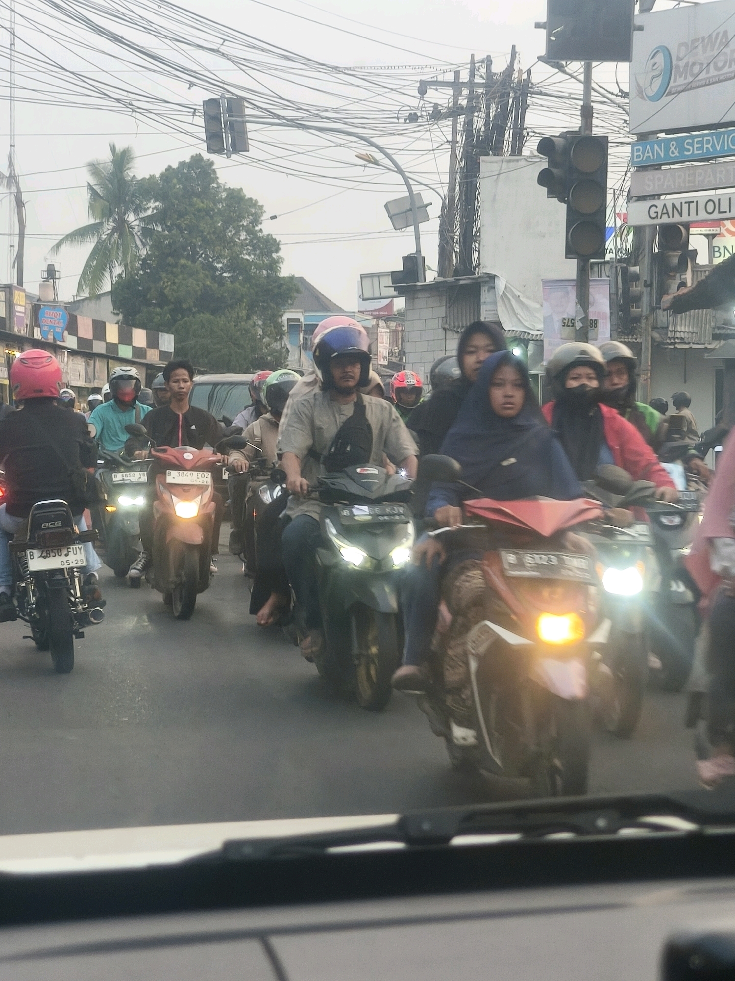 In the city of Karangsatria, Indonesia, a busy street scene unfolds with numerous motorcycles navigating traffic. A group of individuals, predominantly young adults and some children, are riding scooters and motorbikes. Several people are wearing helmets in various colors, including pink, blue, green, and black. Some riders are also wearing backpacks and casual clothing. The environment is characterized by an overhead network of electrical wires and poles, typical of urban Indonesian streetscapes. Traffic signs are visible in the background, including "DEWA MOTOR" and "GANTI OLI." The road surface appears to be asphalt. The lighting suggests it is daytime, possibly overcast, as indicated by the diffused light and lack of strong shadows. The image captures a moment of everyday life in a bustling Indonesian city, with a focus on the prevalent mode of transportation.