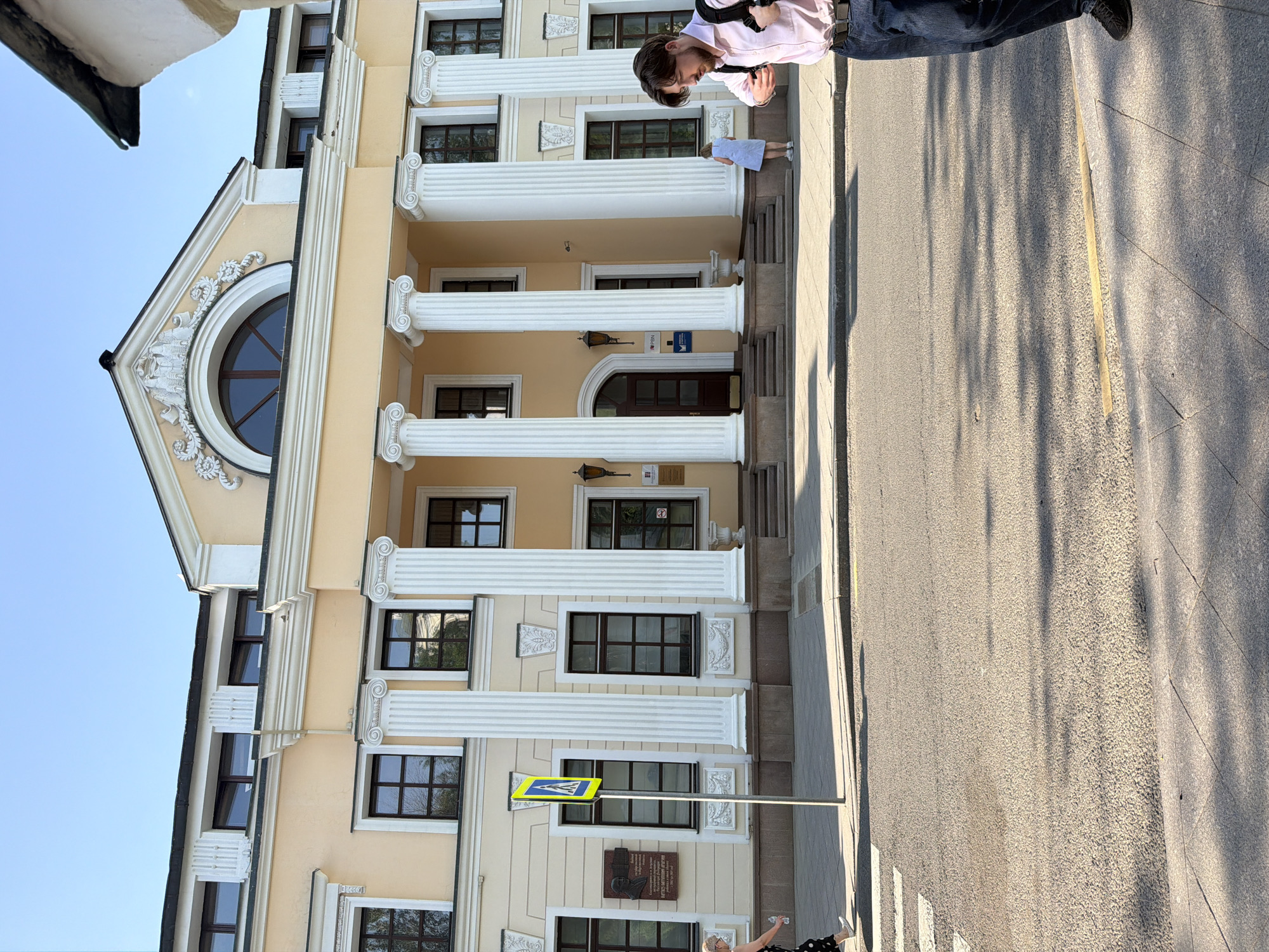 The image shows the exterior of a building with classical architectural elements, including columns and a pediment with a round window. The building's facade is painted in a light yellow color with white trim. A pedestrian crosswalk and a street sign are visible in front of the building. Two people are present: one man stands on the steps leading to the building's entrance, and another individual stands on the sidewalk near the crosswalk. The location is identified as Moscow, Russia.
