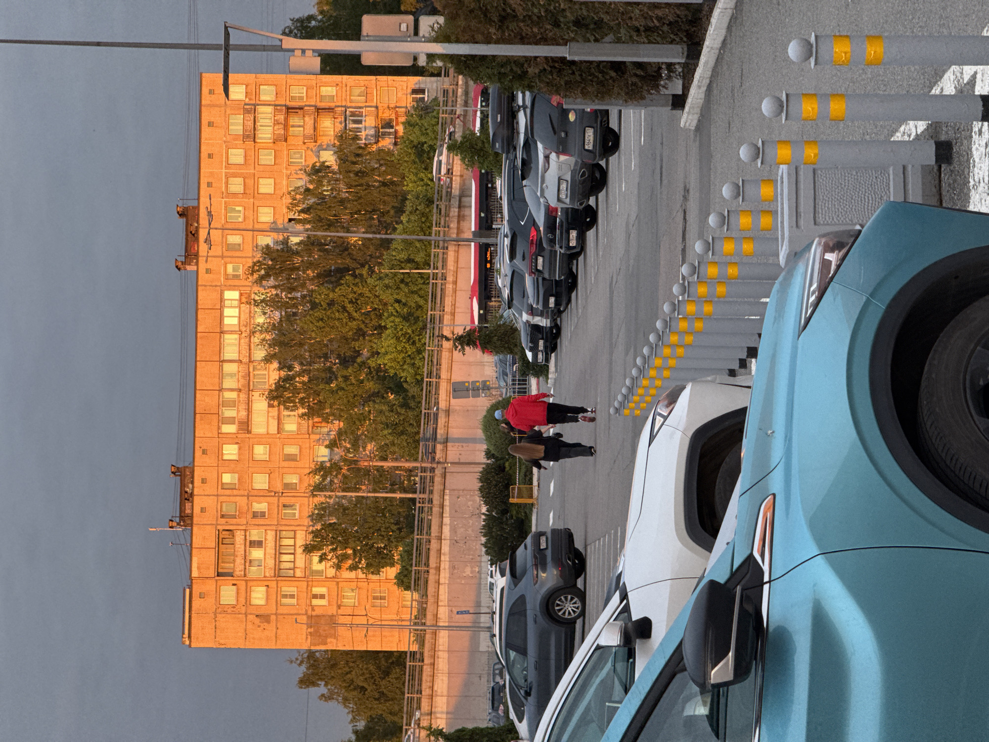 The image depicts an outdoor urban scene, likely a street or parking area adjacent to a large building, captured during late afternoon or early evening. The sky overhead is a muted grey-blue.
On the left side of the frame, a tall, multi-story building with numerous square windows is prominently featured. Its facade is bathed in a warm, golden light from the setting or rising sun, contrasting with the cooler tones of the sky. Green trees are interspersed between the building and the paved area below.
In the middle and foreground, a parking lot contains several parked cars, mostly dark-colored SUVs or sedans. Some vehicles have roof racks or cargo boxes. A light blue car is partially visible in the immediate foreground on the right. Two individuals are walking on the paved surface near the parked cars. One person wears a red jacket and dark pants, while the other wears a dark jacket and pants, both appearing to carry bags.
A row of grey bollards with yellow reflective bands lines the right edge of the parking area, separating it from an adjacent section. Utility poles and power lines are also visible against the sky.
Visible Russian license plates on some cars, such as "P944YH 178 RUS" and "K337YM 178", confirm the location as Saint Petersburg, Russia. The scene captures a moment of daily urban life, with no specific emotional cues evident.