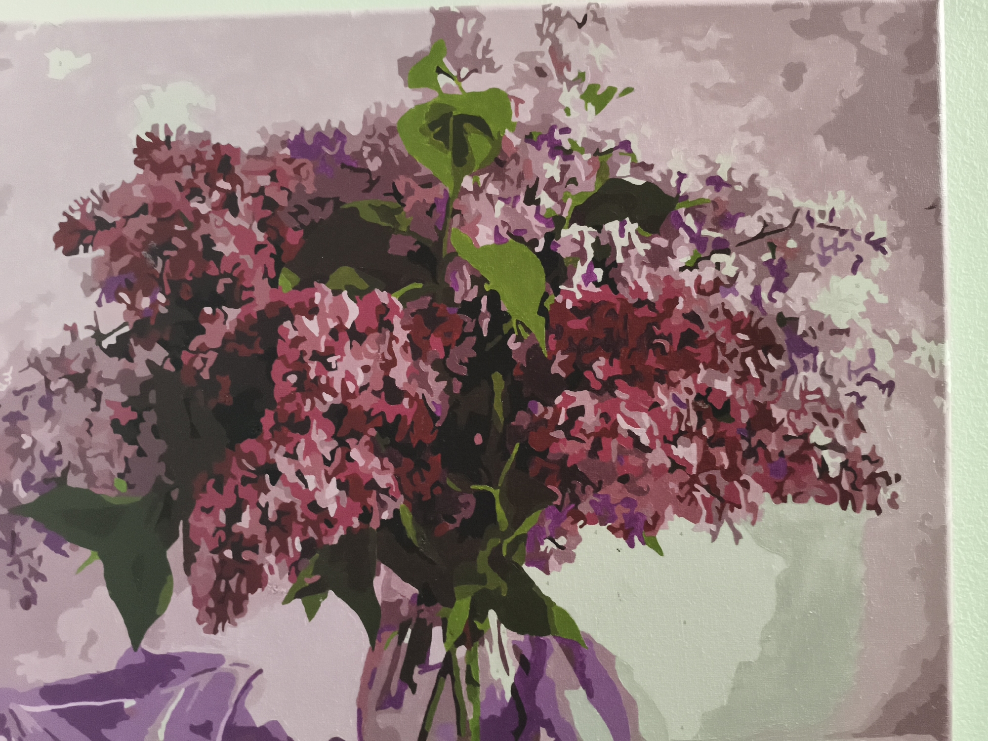 The image shows a painting of a lilac bouquet in a vase. The flowers are predominantly pink and purple, with some green leaves visible. The background is a muted, pale purple color. The vase is a light purple color. The style of the painting is simple and slightly abstract. There is no information to determine the location in Nowy Tomyśl, Poland.
