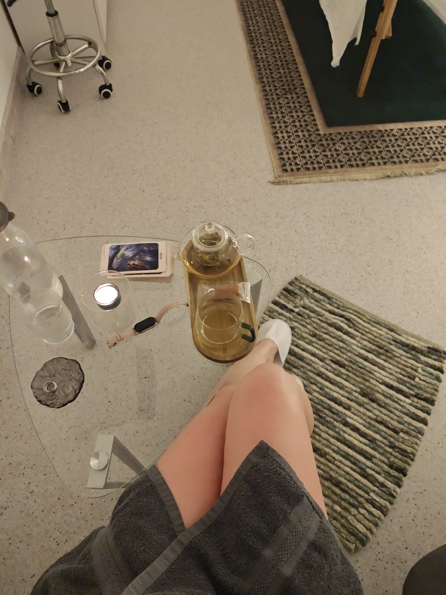 The scene is an indoor setting, possibly a spa or wellness center. A person is seated with their legs crossed, draped in a gray towel. A small, round glass table is next to them, holding a teapot, a cup of tea, a bottle of water, a lit candle, a stack of cards, and a smartwatch. On the floor are two small rugs, one striped and the other with a textured border. In the background, there's a rolling stool and a larger rug under a table. The city of Vinnytsia, Ukraine, cannot be confirmed as the location based on this image alone.