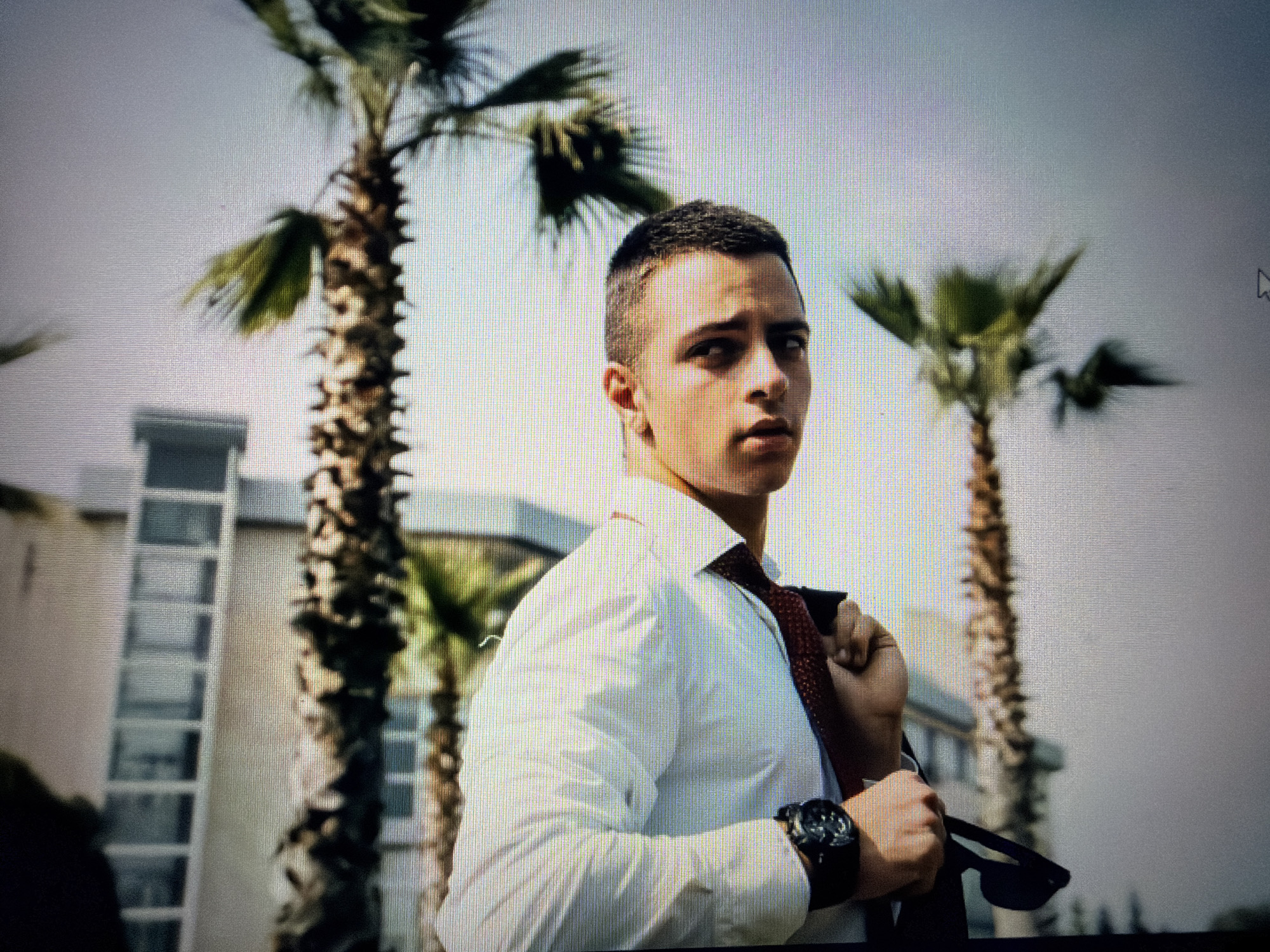 The image depicts a young man outdoors in an urban environment, potentially in the city of Unknown, Unknown. He is wearing a white collared shirt and a dark tie, which he is adjusting with his hand. He has a dark watch on his left wrist. He appears to be looking directly at the camera.
In the background, there are palm trees and a building with multiple windows. The sky is overcast. The lighting suggests it is daytime.