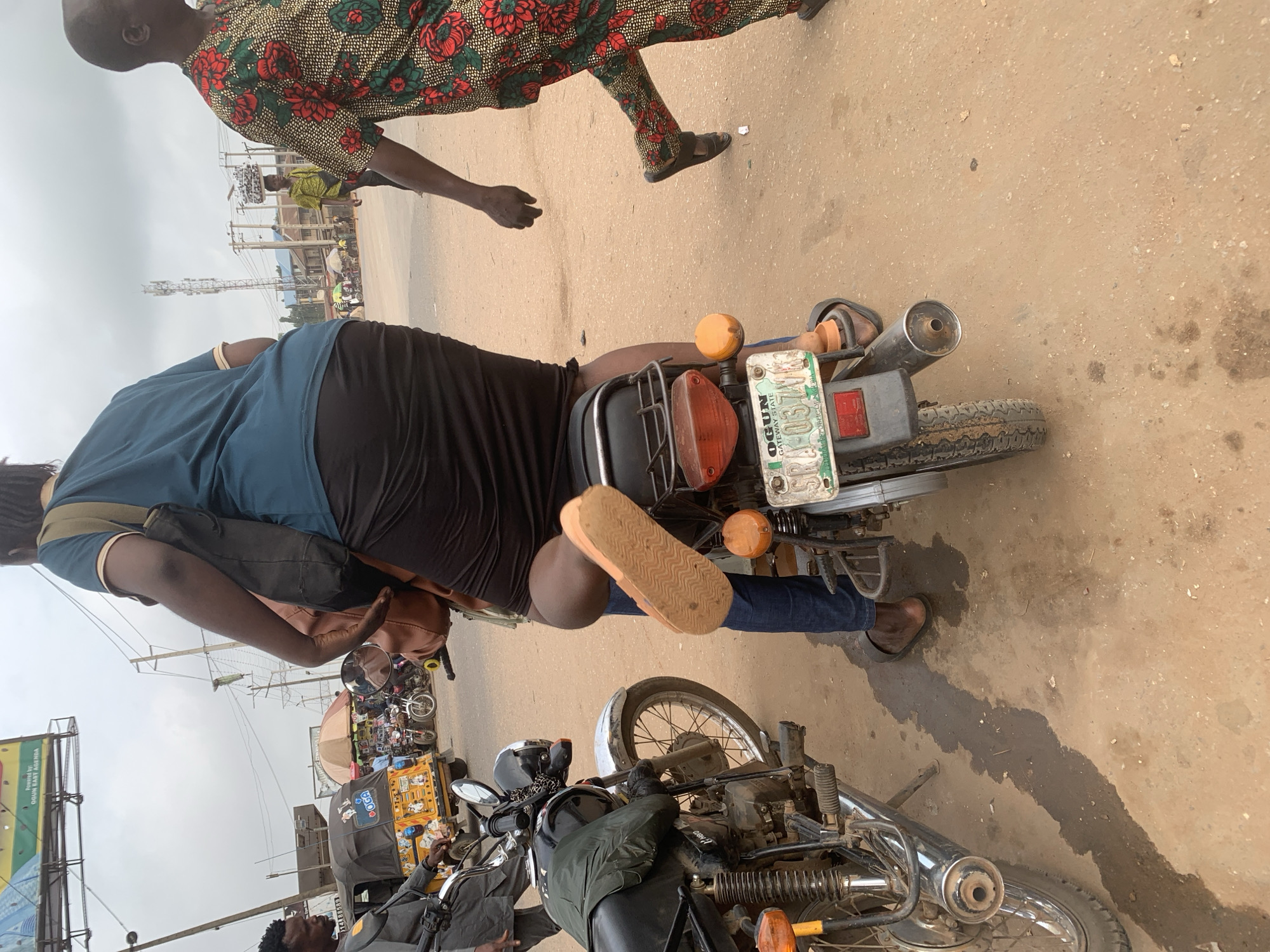 The image depicts a street scene in Shagamu, Nigeria, likely on a dusty, unpaved road. In the foreground, a person is seated on the back of a motorcycle, their legs visible. The motorcycle is black and appears to be a common model in the region. To the left, another motorcycle is parked, also black, with a rider wearing a dark jacket. In the background, a yellow trishaw with "DOGA" written on it is visible, suggesting a commercial area. Further in the background, other motorcycles and people are present, along with some market stalls under umbrellas. A man in a colorful patterned shirt walks past, and a cellular tower is visible in the distance. The sky is overcast, indicating a cloudy day. The overall impression is of a busy, everyday street scene in a Nigerian city.