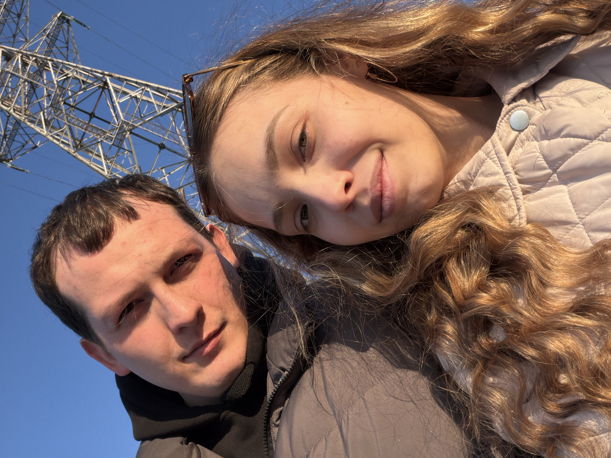 A selfie shows a man and a woman standing close together in an outdoor setting. The woman, with long wavy blonde hair, is positioned on the right and leans her head towards the man. She is wearing a light-colored quilted jacket with a button. The man, with short dark hair, is on the left and is wearing a dark hoodie under a grey jacket.
In the background, an electricity pylon stands against a clear blue sky. This suggests the location might be near infrastructure in Chaykovsky, Russia.