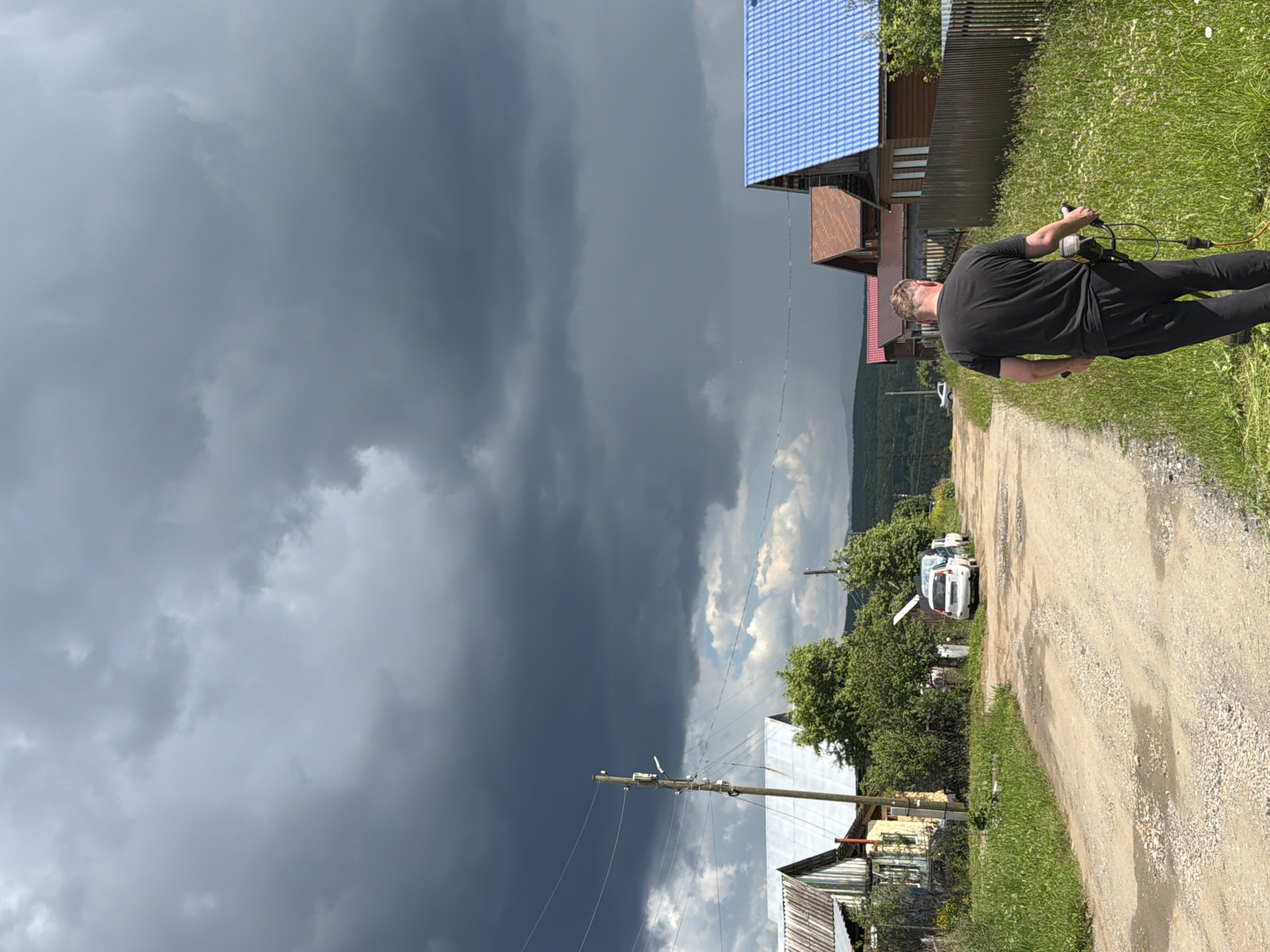 A man is seen from behind, walking down a dirt road in Mezhevoy, Russia, operating a weed trimmer. He is wearing a black t-shirt and black pants. The road is lined with grass and houses, some with blue and brown roofs. The sky is filled with dark, ominous clouds, suggesting an approaching storm. There are several cars parked along the side of the road. A utility pole with wires is visible on the left side of the road. The overall scene depicts a rural setting under dramatic weather conditions.