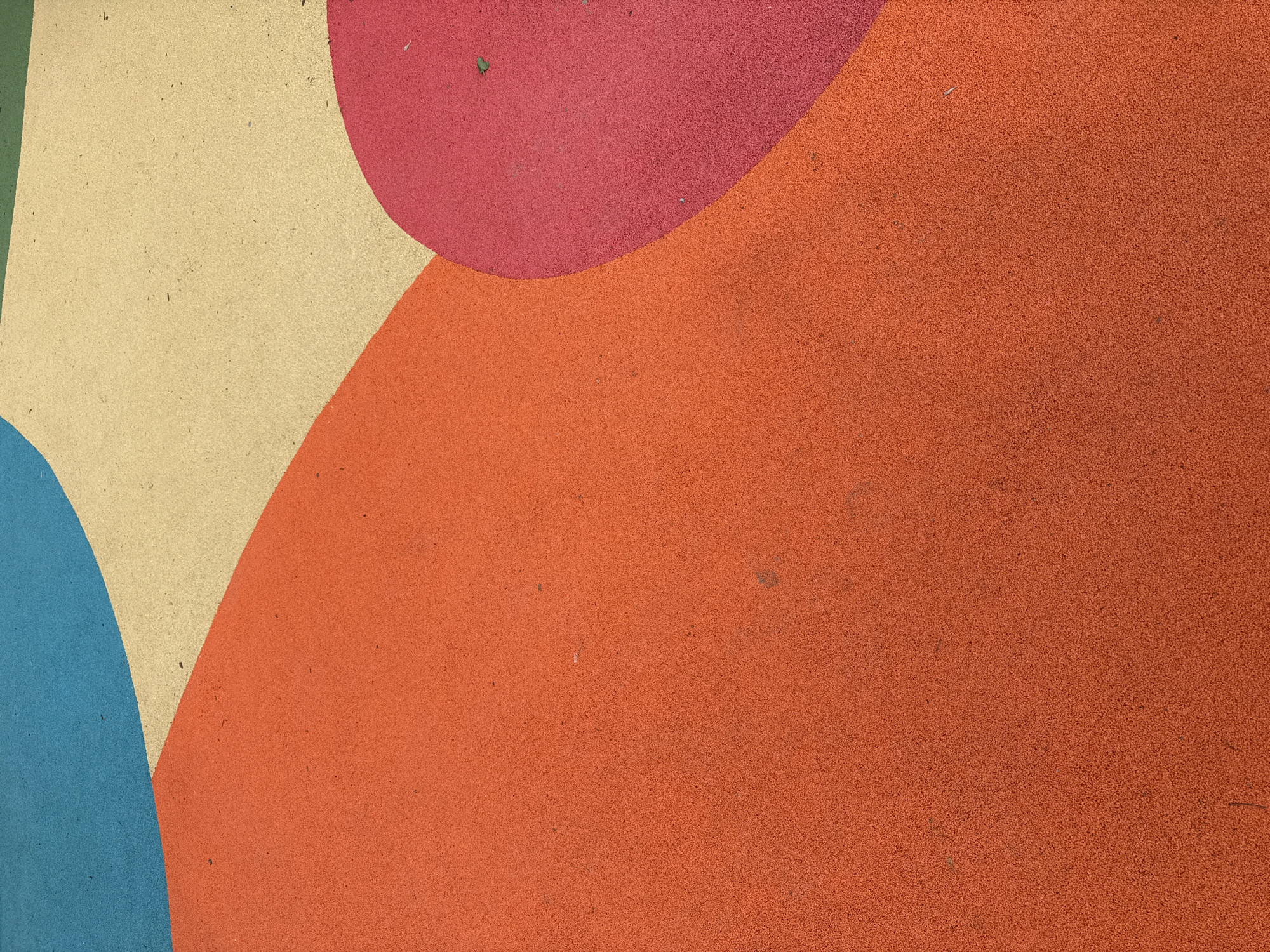The image shows a section of a playground surface in Moscow, Russia. The surface is made of a soft, granular rubber-like material and is divided into several colored sections: blue, beige, red, and orange. The colors are arranged in overlapping, rounded shapes. A single small leaf is visible on the red section. The surface appears slightly worn, with small imperfections and dirt particles scattered throughout.