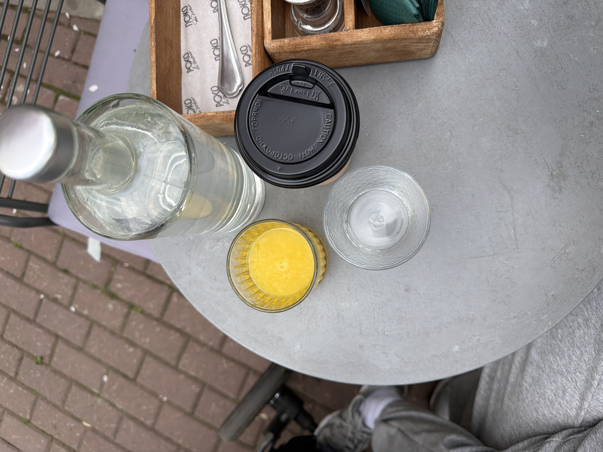 A high-angle shot shows a round, gray outdoor table with several items arranged on it. A glass bottle with a silver cap sits to the left, partially off the table. Next to it is a disposable coffee cup with a black lid. Further to the right is a glass of water and a glass of orange juice. At the top of the frame, there is a wooden container holding napkins and cutlery. The lower right corner of the image shows a person wearing gray clothing. The table is situated outdoors, as evidenced by the view of brick paving stones and hints of greenery in the background. The branding on the napkins indicates a location related to "POKO di buono," potentially a restaurant or cafe. The Cyrillic writing on the coffee cup lid reads "ОСТОРОЖНО! ГОРЯЧО!" (CAUTION! HOT!). Based on the Cyrillic writing, it is likely that the location is in a Russian-speaking region.